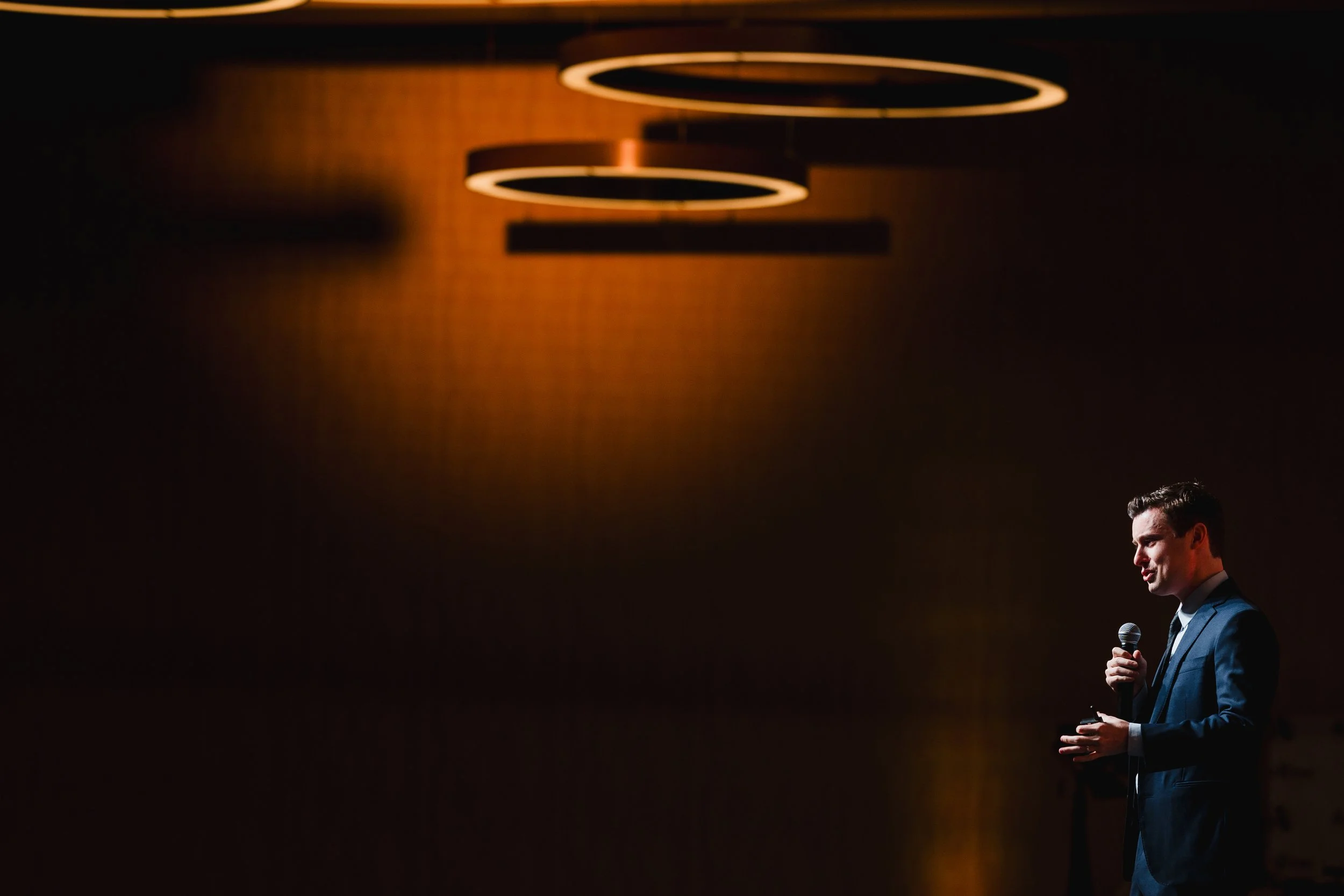 A man in a suit stands on stage holding a microphone, with orange circular ceiling lights overhead, performing or giving a speech in a darkened room.