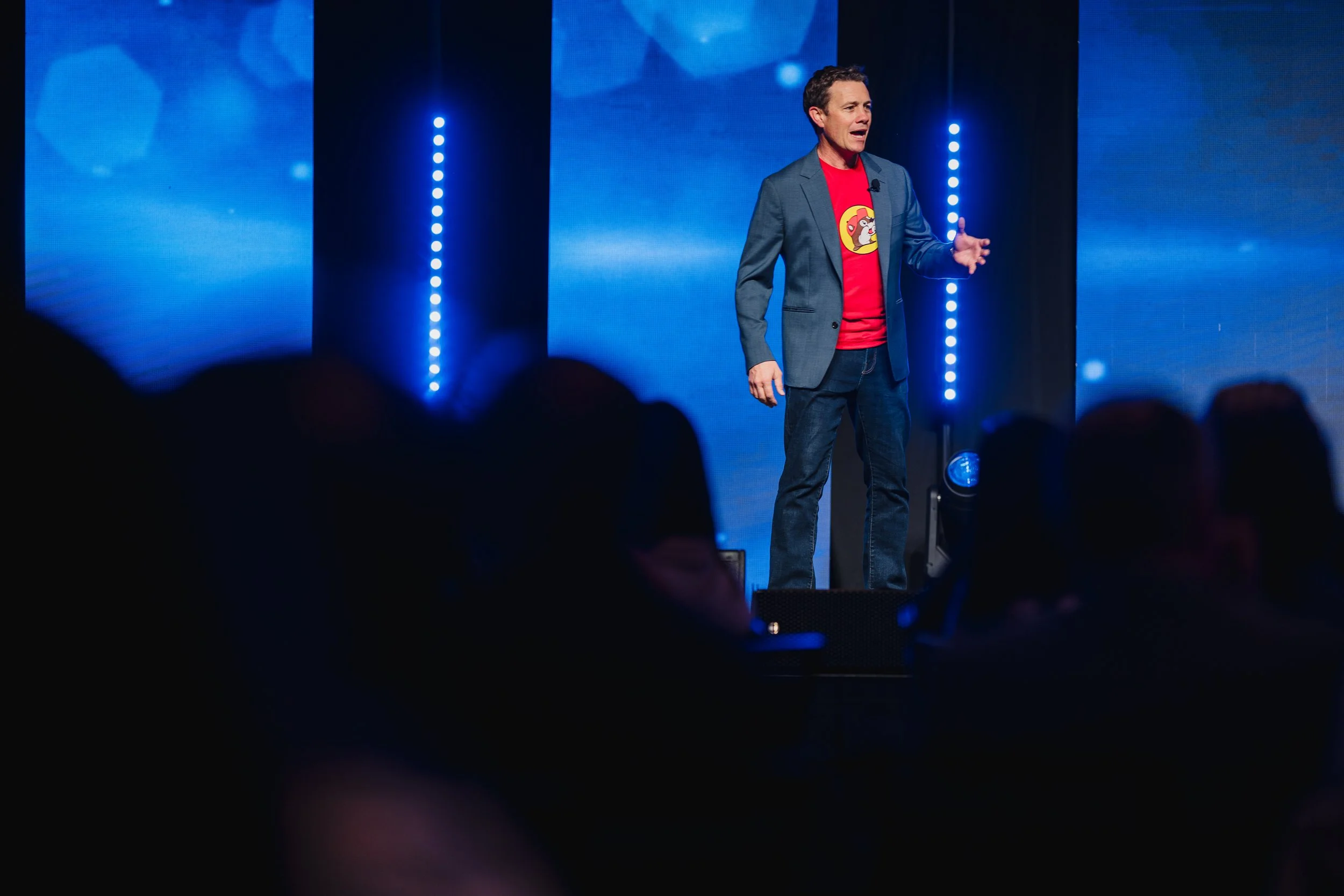 A man in a gray blazer and jeans speaking on stage with a blue background and vertical lights, wearing a red t-shirt with a cartoon character.
