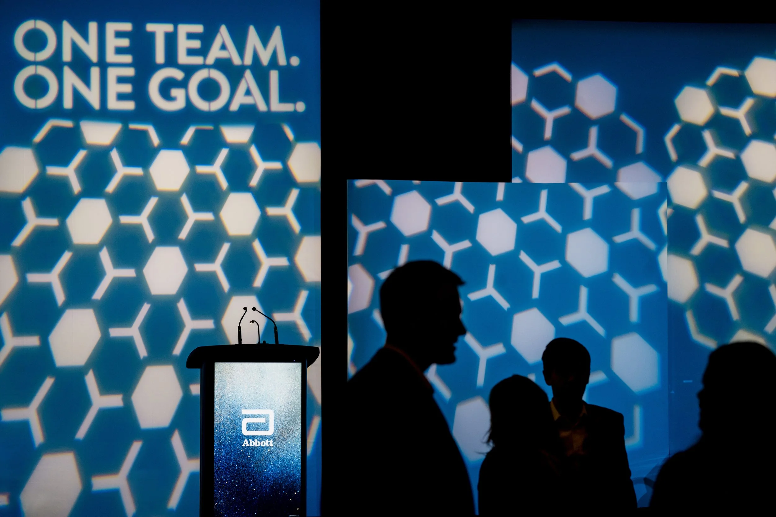 Silhouettes of individuals in front of a large screen displaying the phrase 'ONE TEAM. ONE GOAL.' with a blue hexagonal pattern background. A podium with the Abbott logo is in the foreground.