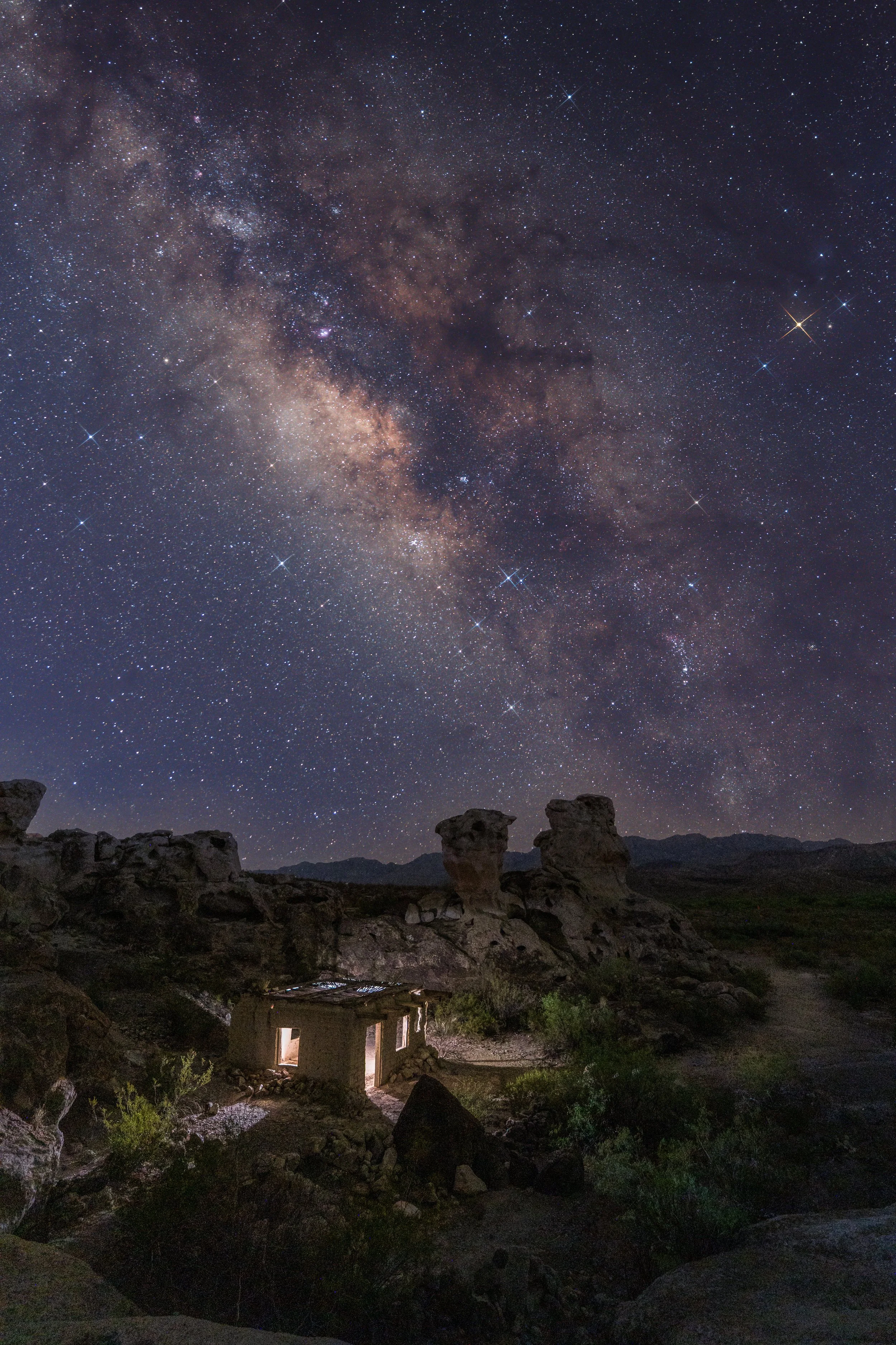 Night sky filled with stars and the Milky Way galaxy over a small house in a rocky desert landscape.
