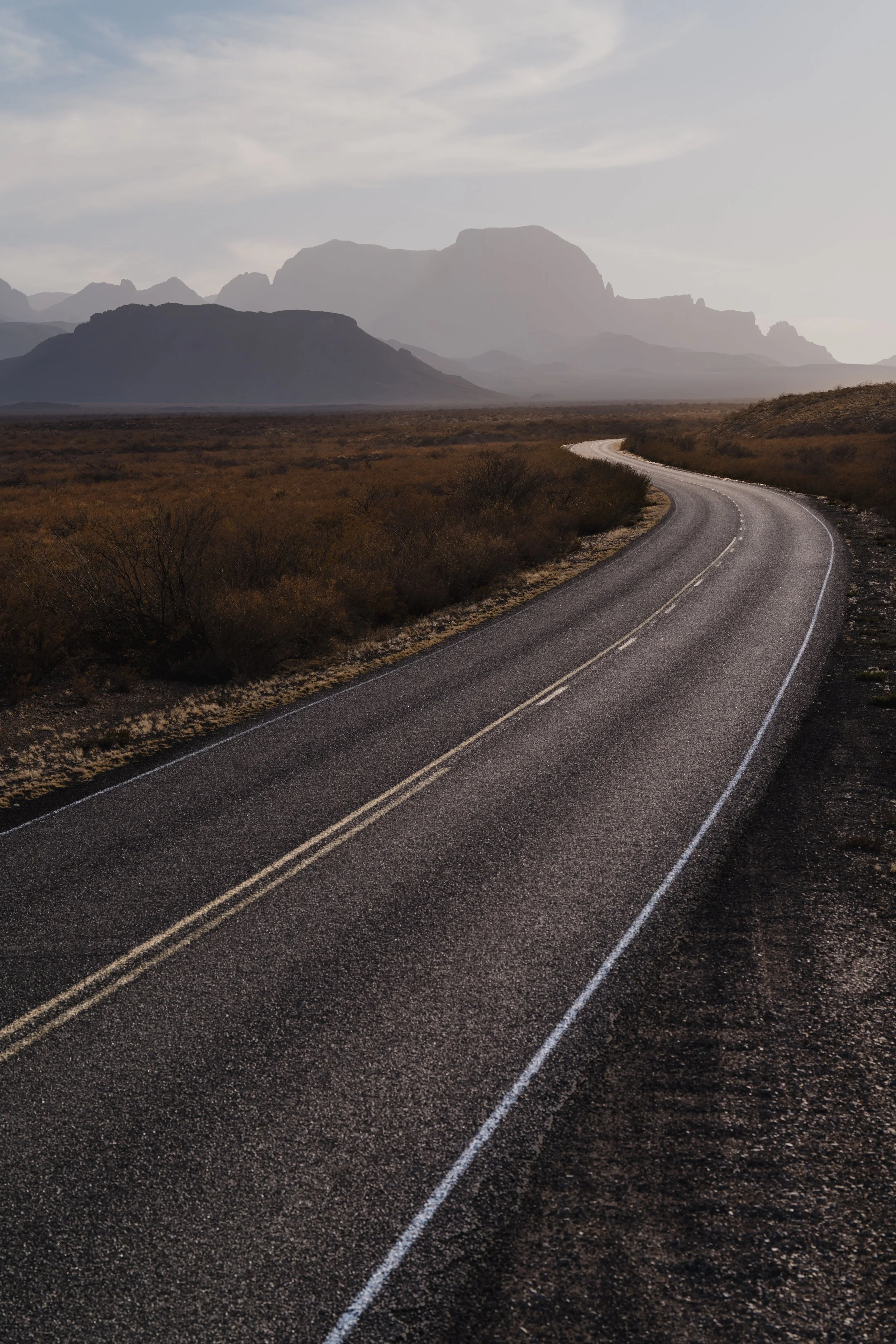 A winding desert road with mountain ranges in the background under a cloudy sky.