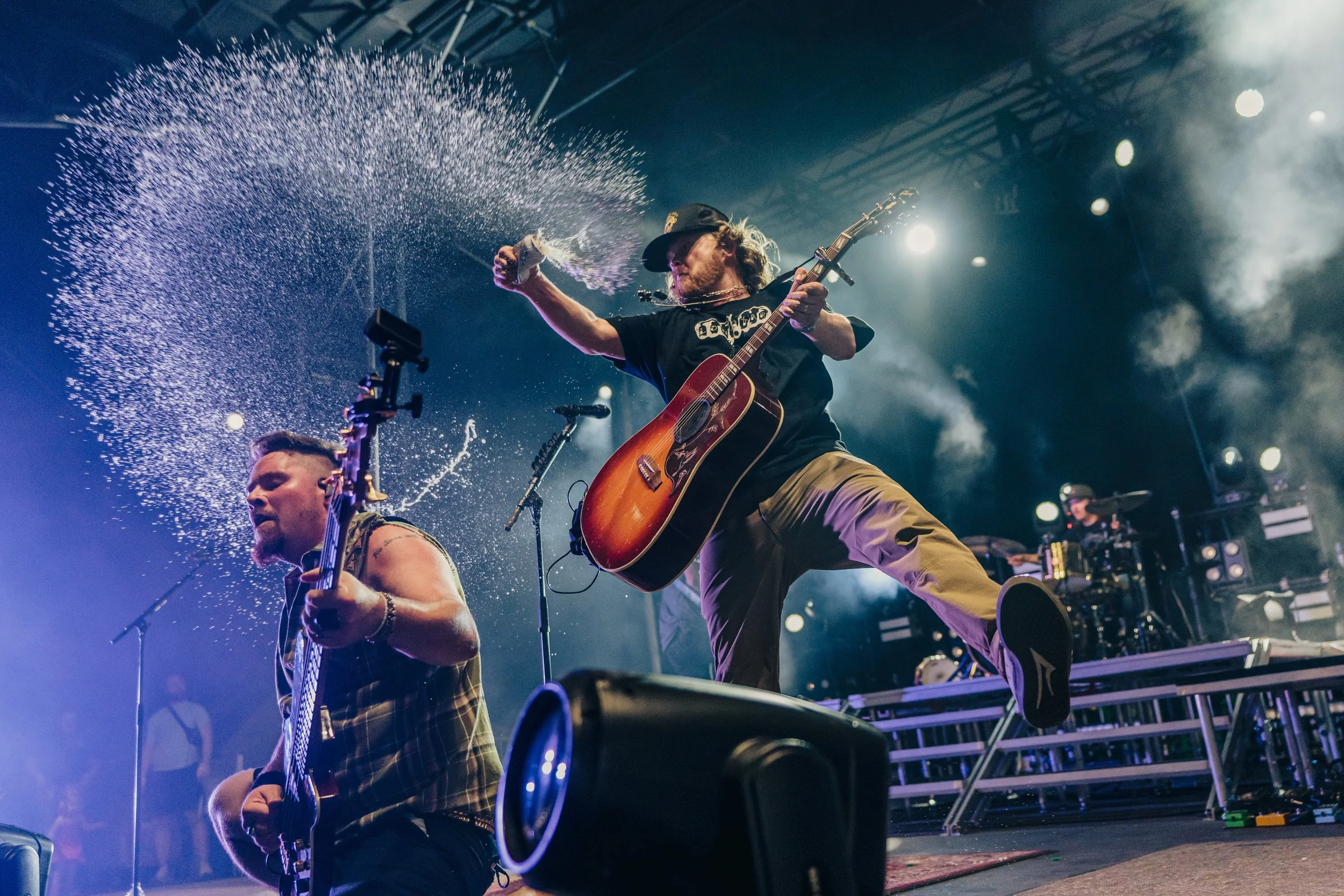 Two musicians performing on stage at a concert, one with an acoustic guitar and the other with an electric guitar, while water is being splashed during the energetic performance with stage lights illuminating the scene.