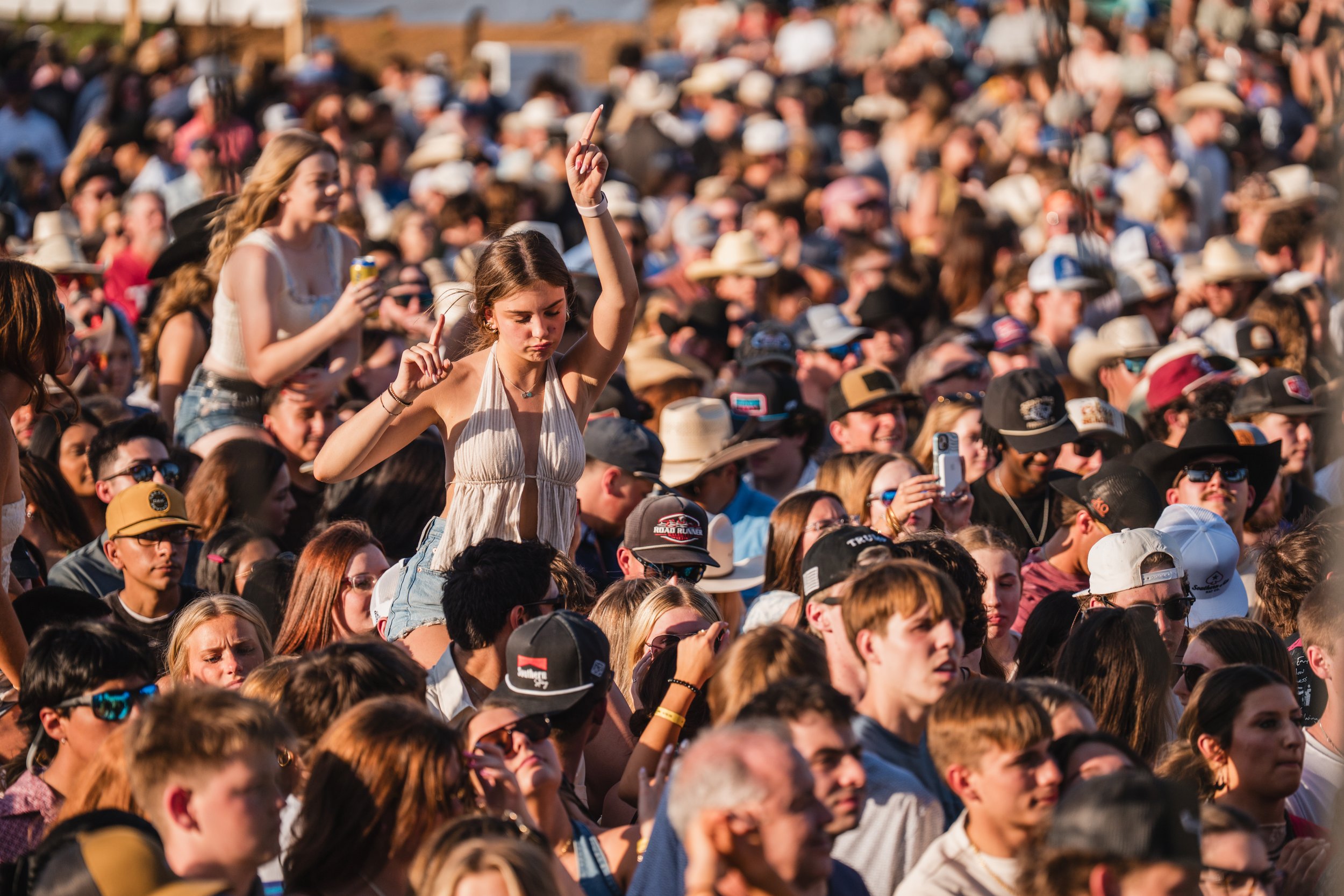A large crowd at an outdoor concert or festival, with a young woman standing on someone's shoulders dancing, surrounded by many people wearing sunglasses and hats.