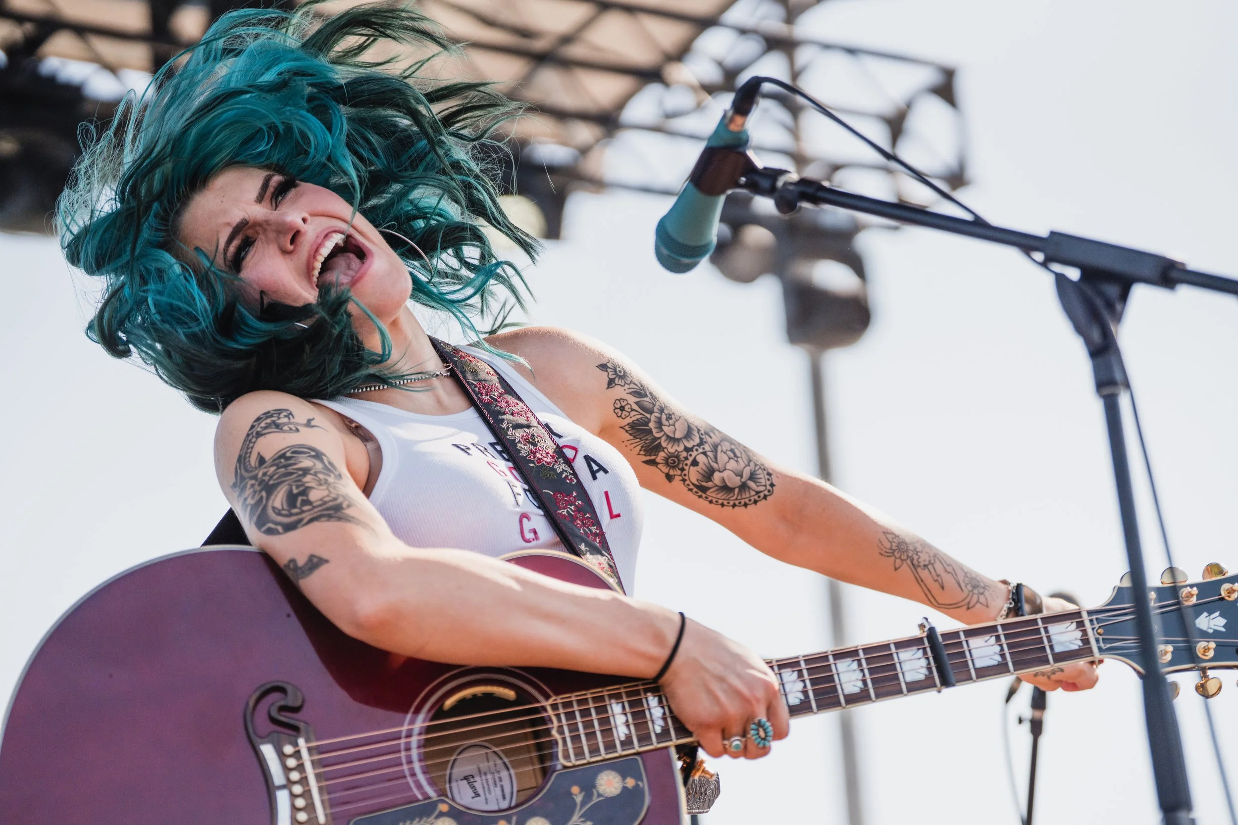A female musician with blue hair and tattoos passionately singing and playing an acoustic guitar on stage.