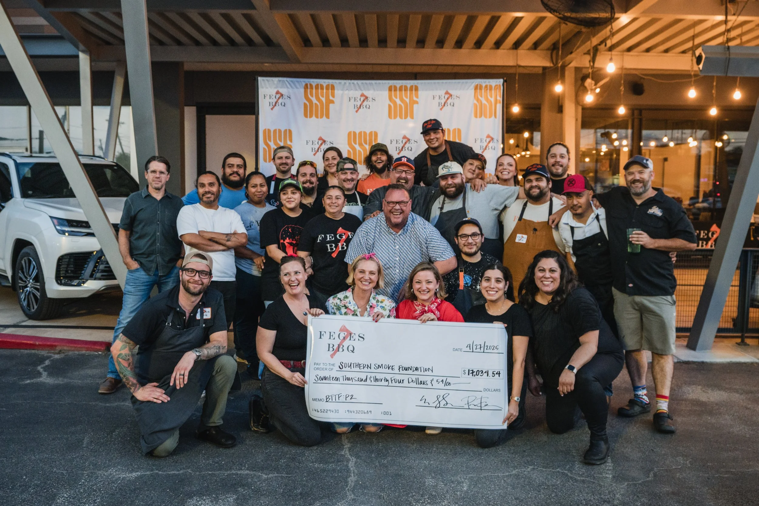 Group of people posing outdoors in front of a barbecue restaurant, holding a large check for over $17,000, celebrating a fundraiser event.