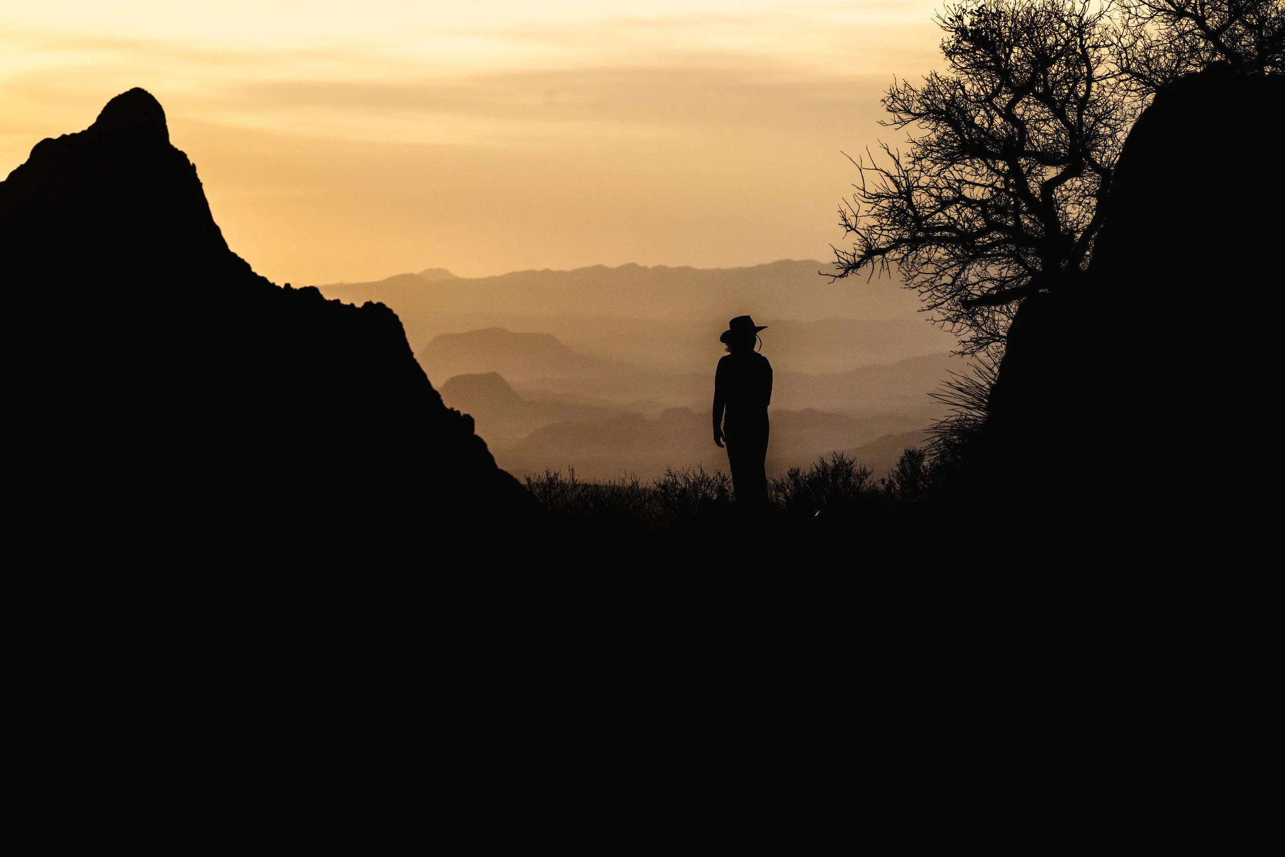 Silhouette of a woman wearing a hat standing between two rocks and trees, with a layered mountain landscape and a sunset sky in the background.
