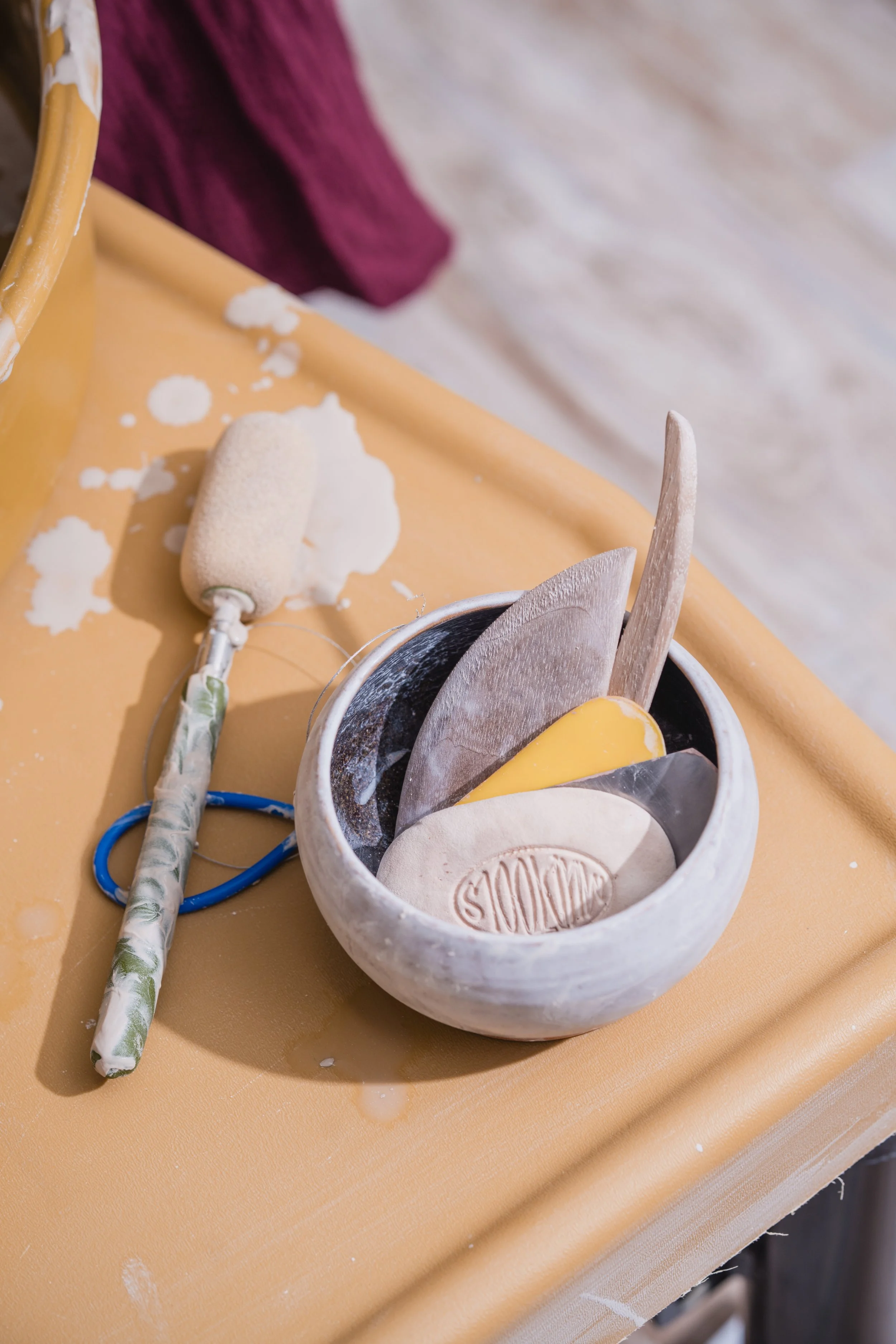 A pottery workspace with a bowl of clay tools and a clay rolling pin on a yellow table.