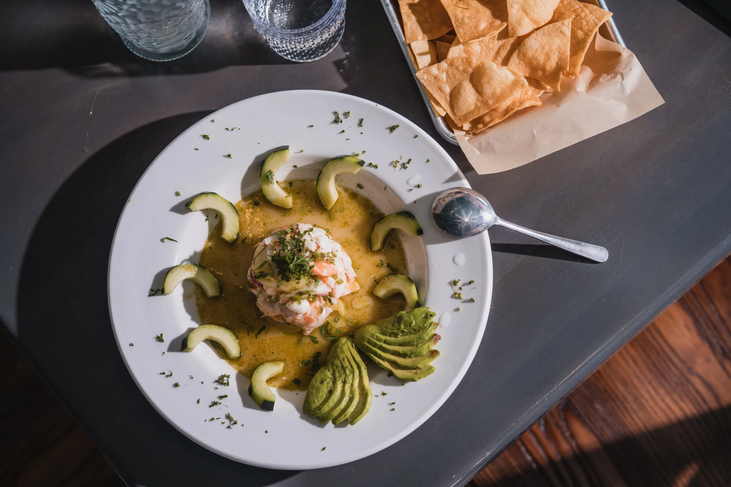 A plate of ceviche with sliced avocado, cucumber, and garnished with herbs, served with lemon or lime juice. There are two glasses of water and a basket of tortilla chips on the dark table.