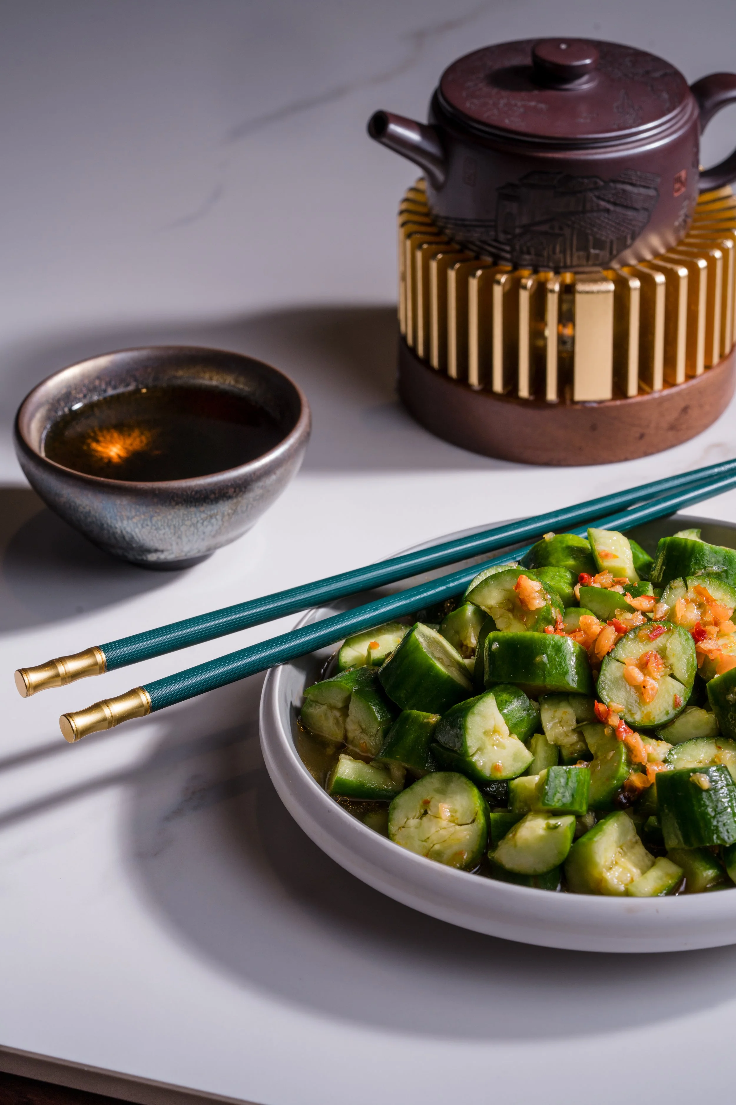 A bowl of chopped cucumbers with a chili sauce on top, Japanese chopsticks, a bowl of soy sauce, and a teapot in the background on a white marble surface.