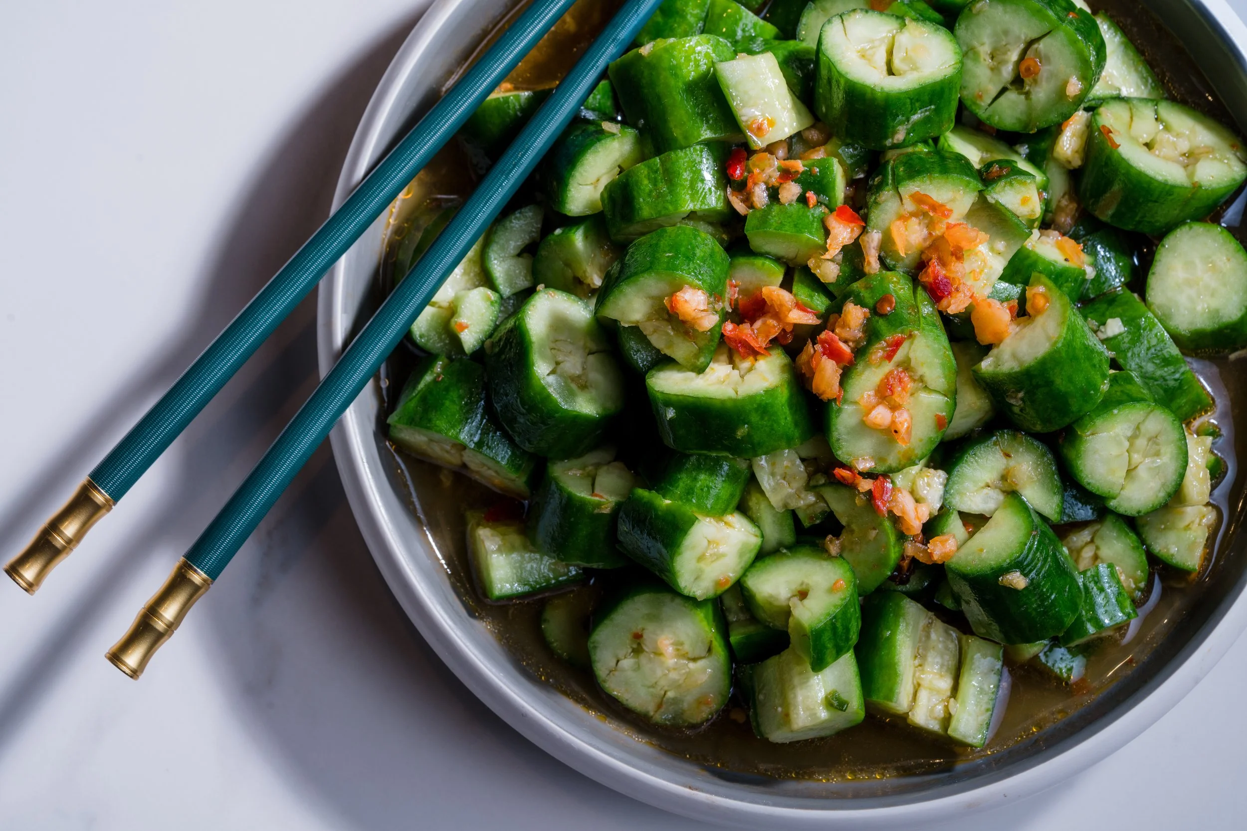 A bowl of chopped cucumbers with garlic and chili flakes, garnished with a liquid sauce, and a pair of teal chopsticks resting on the side.