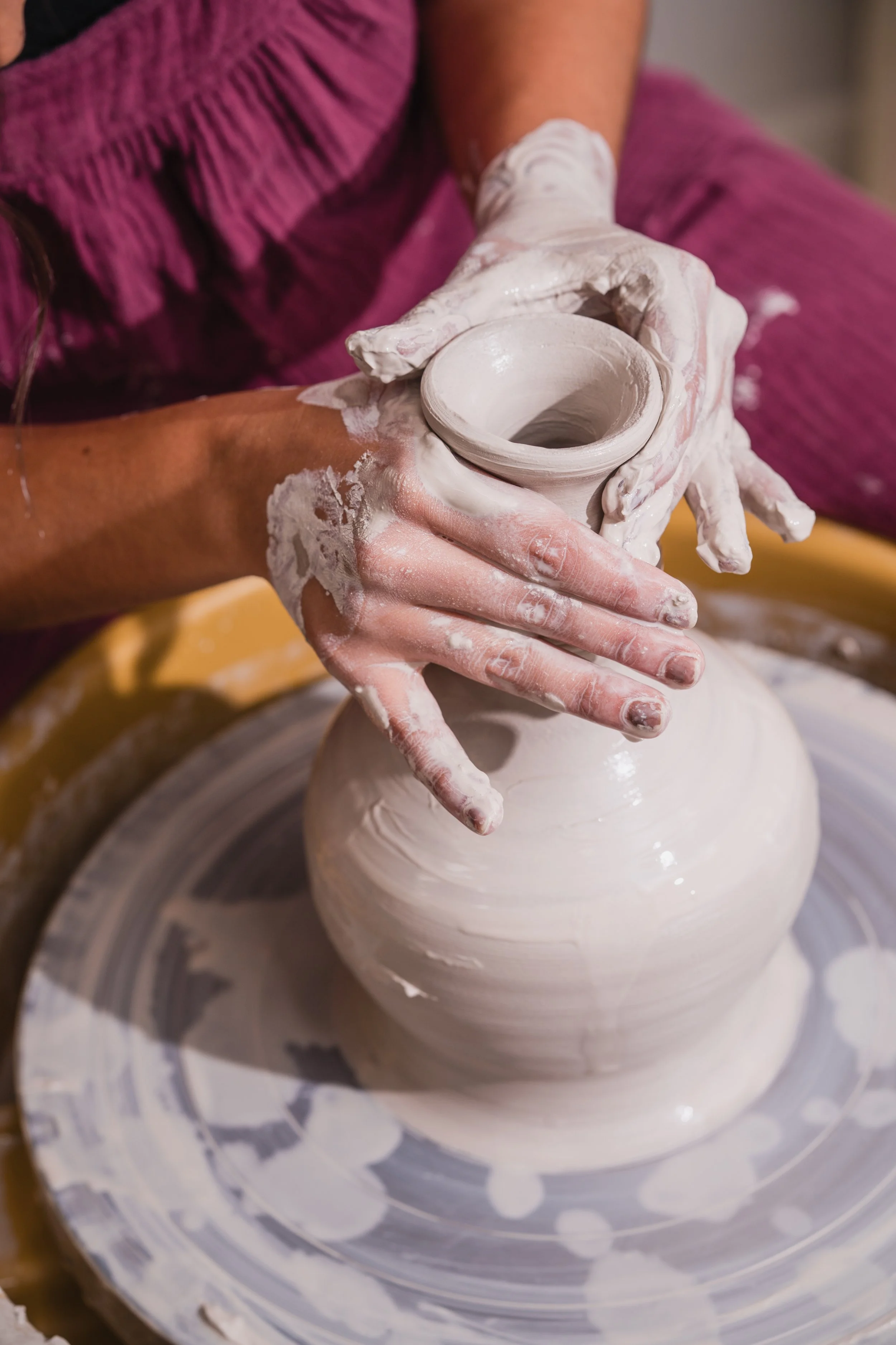 Person shaping a ceramic vase on a pottery wheel with white clay, wearing a purple top, hands covered in clay.
