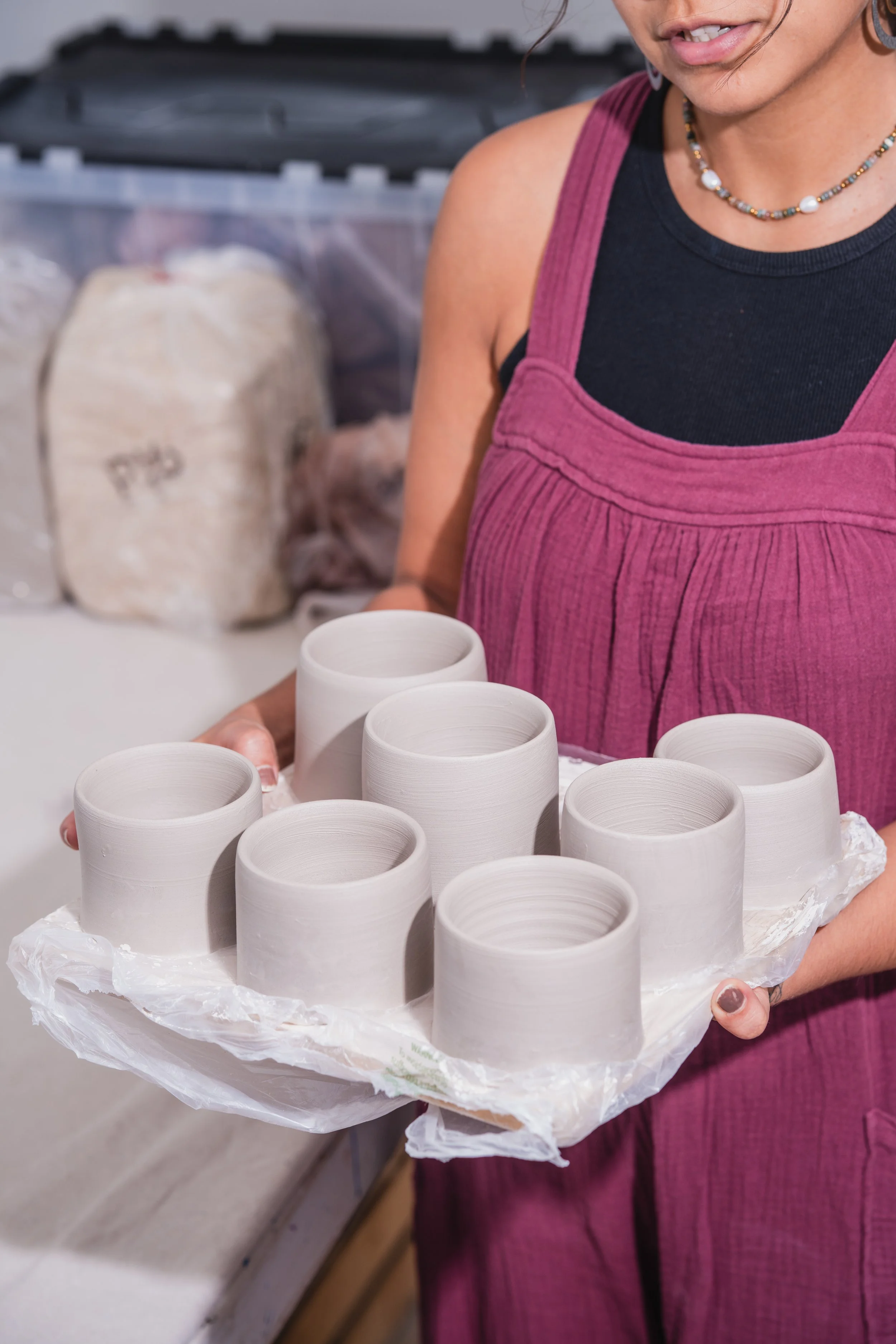 A woman holding a tray of eight unfinished ceramic cups in a pottery studio.