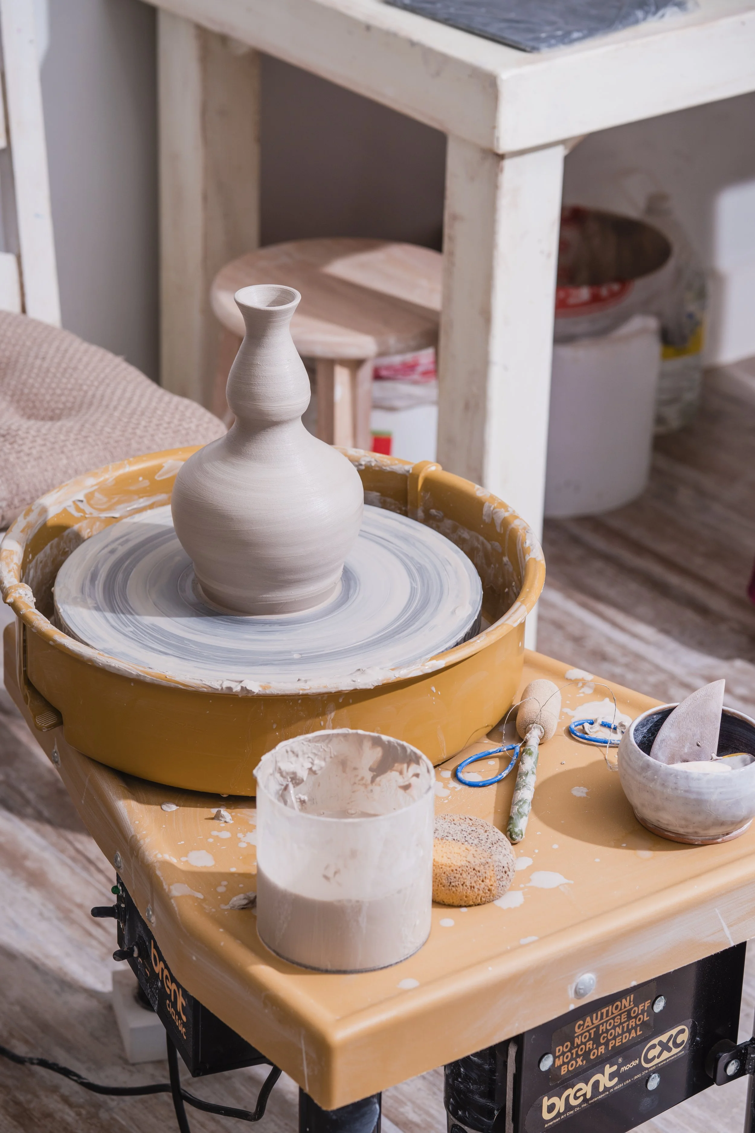 A pottery wheel with a partially finished clay vase in a ceramics studio. Various pottery tools and supplies are on the table nearby.