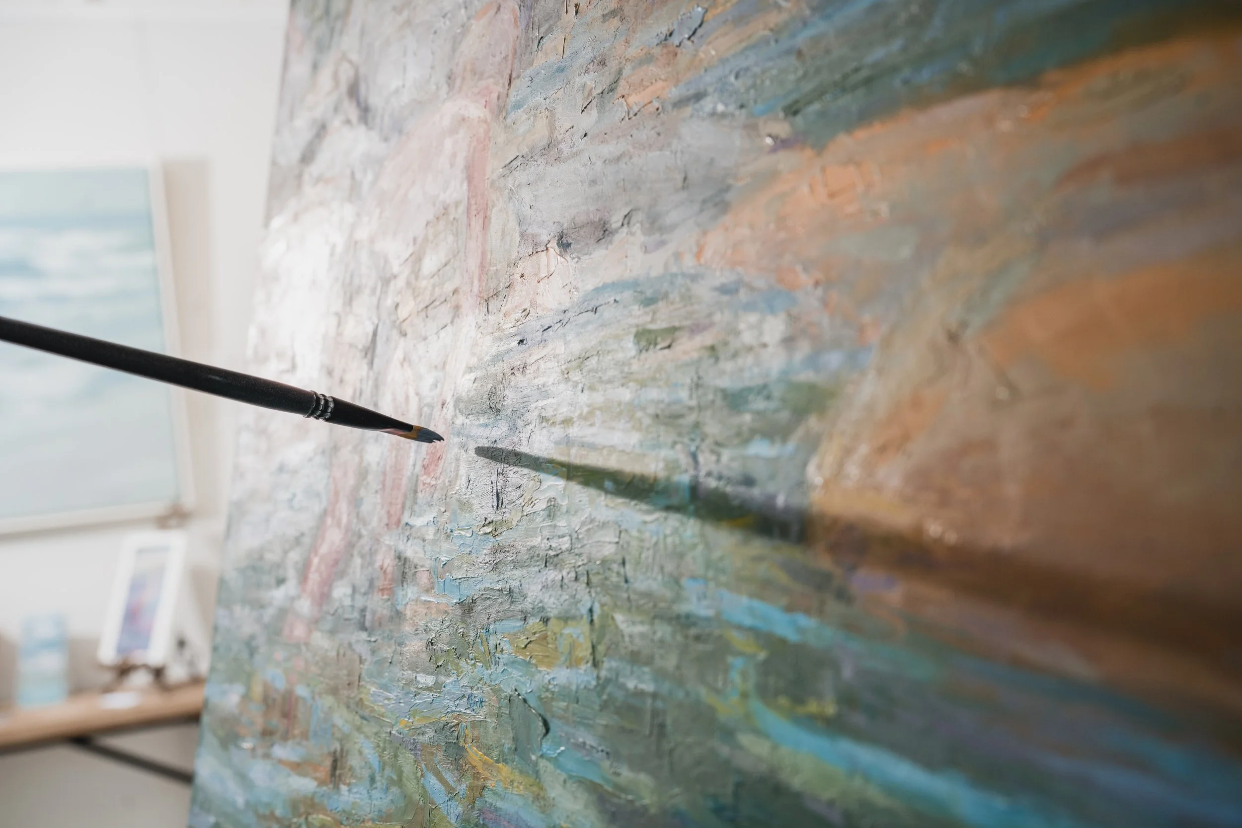 Close-up of an artist's brush touching a textured, abstract oil painting with shades of blue, green, brown, and white on a canvas.