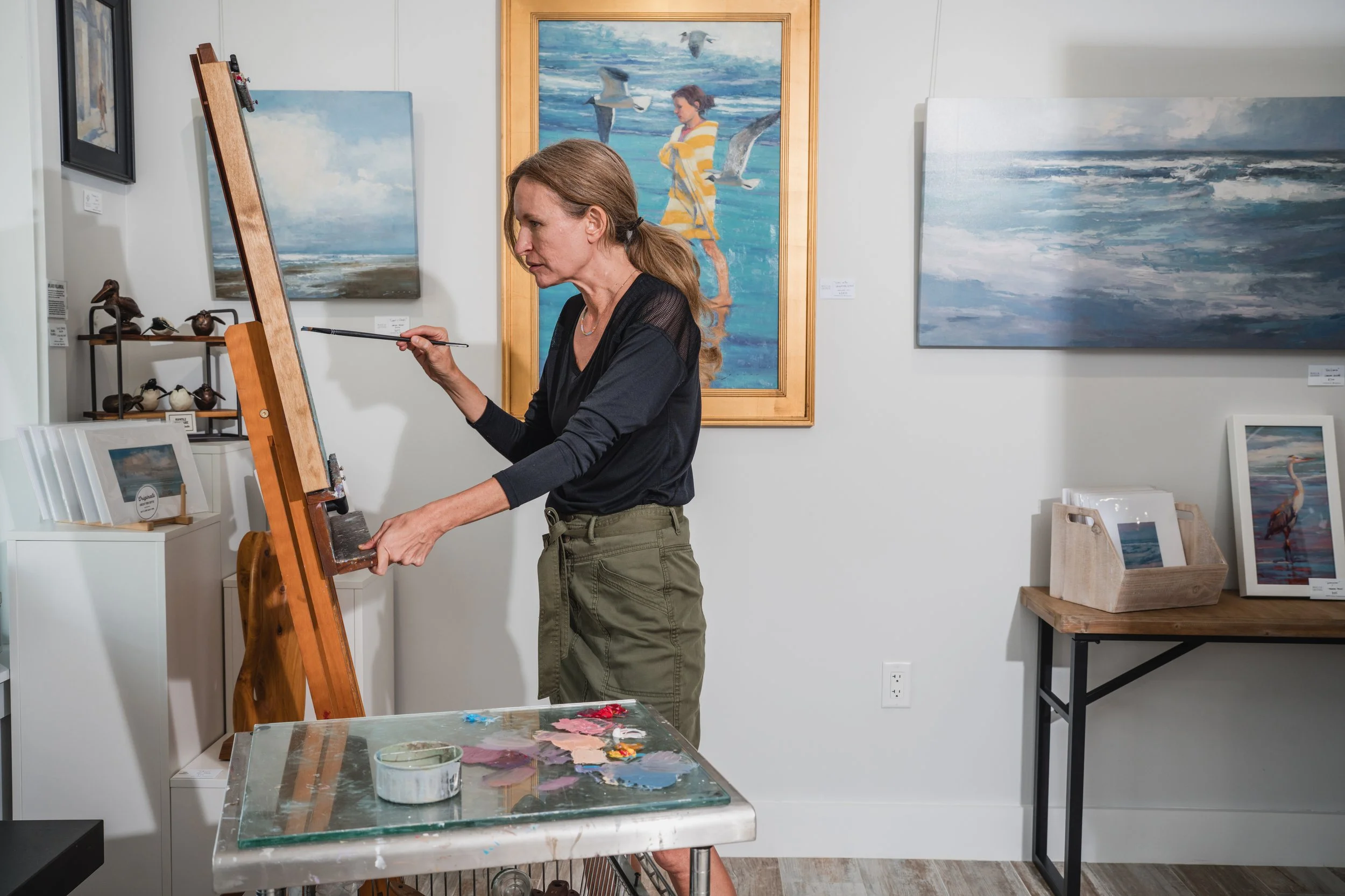 A woman painting on an easel inside an art gallery, surrounded by ocean-themed artwork and decorative bird sculptures.