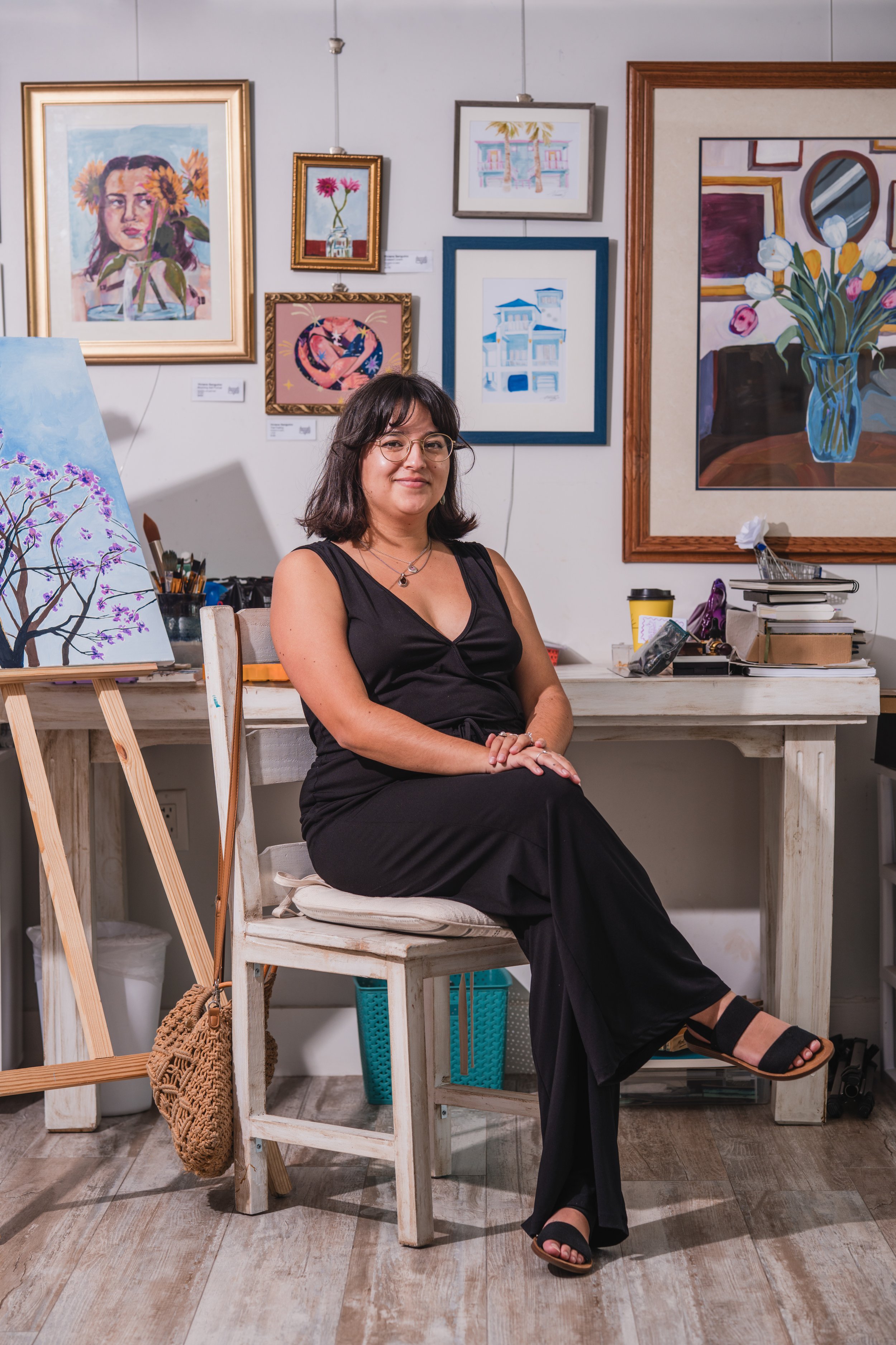 A woman wearing a black sleeveless dress and glasses sitting on a wooden chair in an art studio, surrounded by colorful paintings and art supplies.