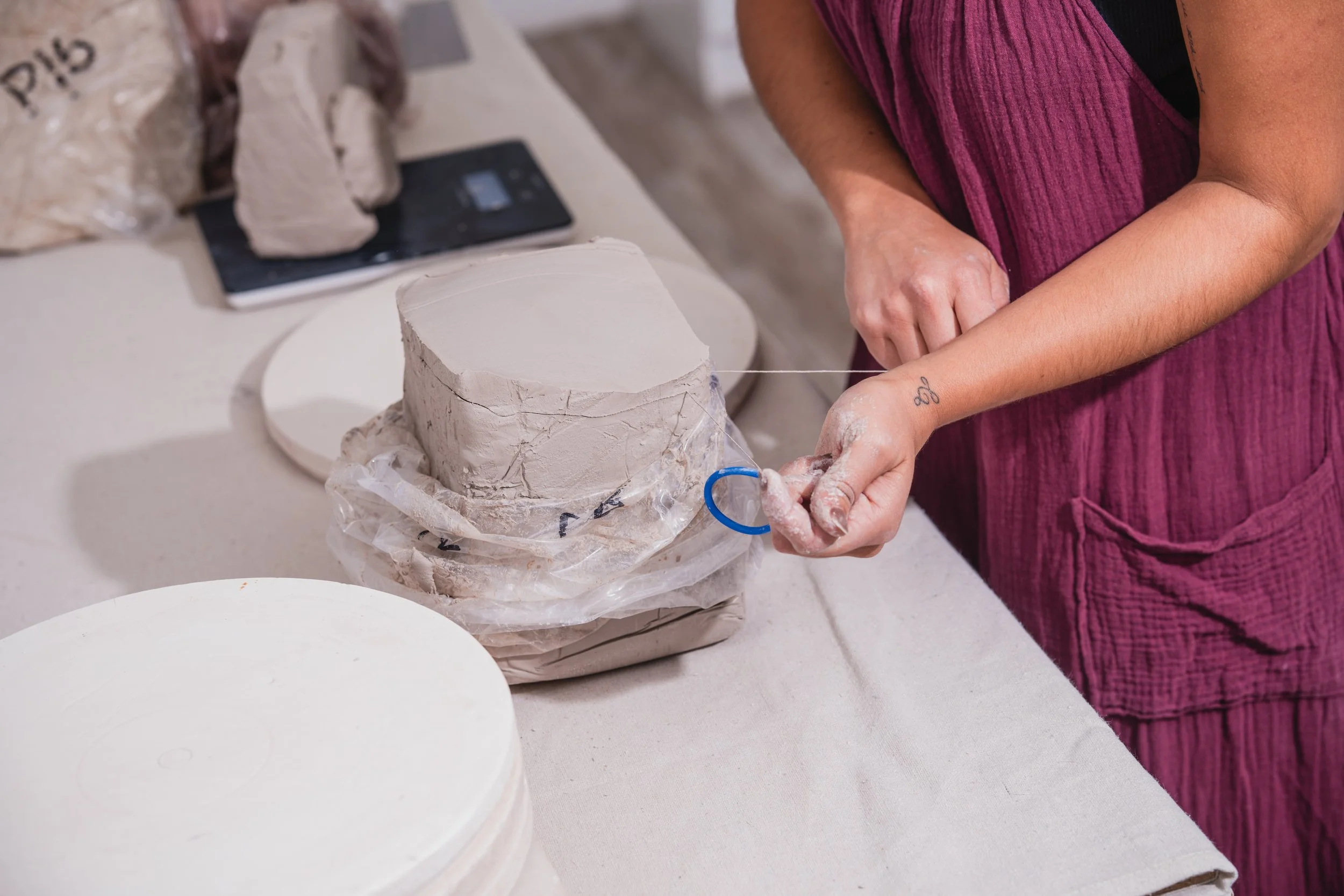 A person in a magenta dress shaping a ceramic piece on a pottery wheel with tools, surrounded by pottery supplies on a table.