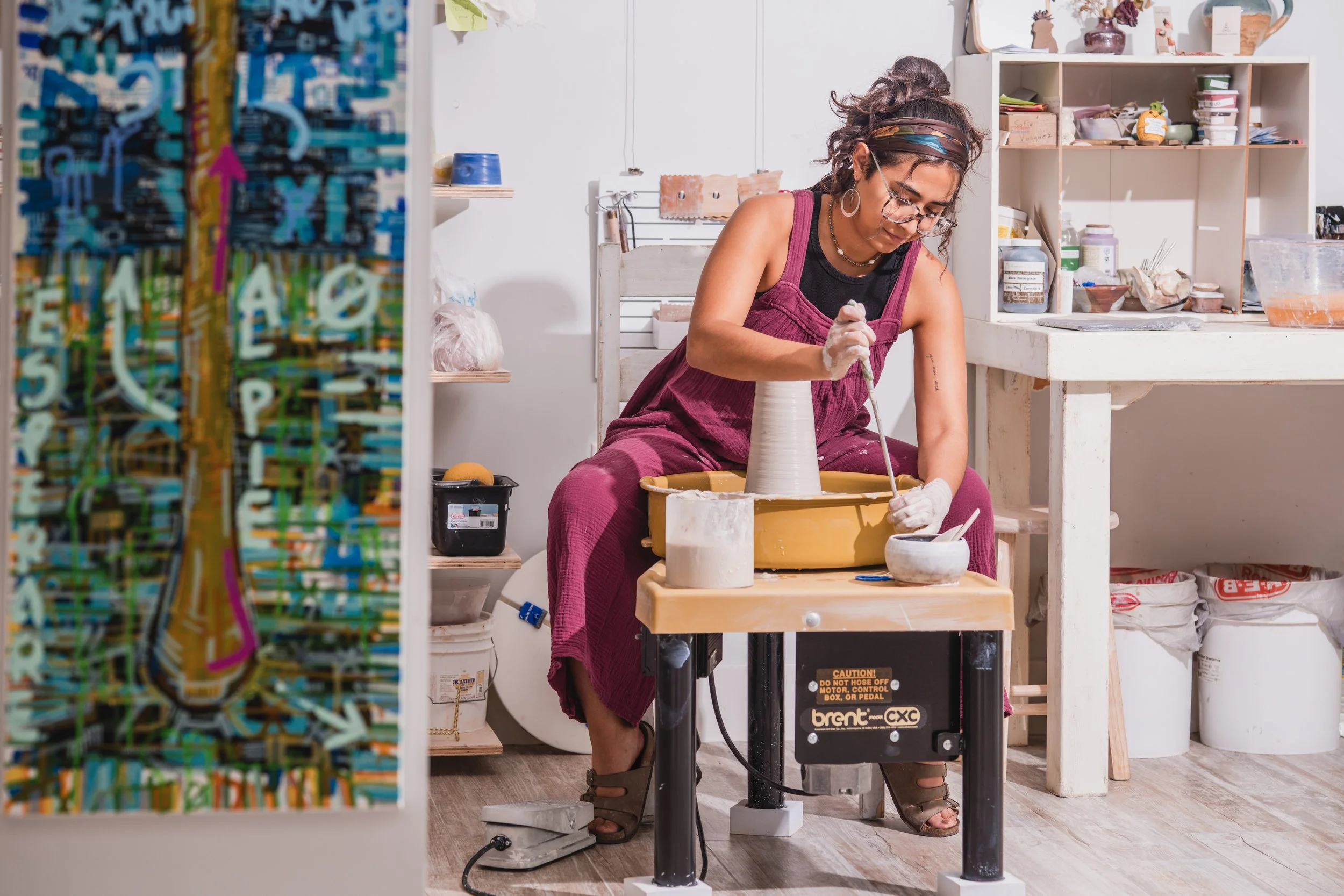 A woman working on a pottery wheel, shaping a clay vase in her pottery studio. She is wearing glasses, a headband, and a sleeveless top, focused on her craft with shelves of supplies and colorful artwork in the background.