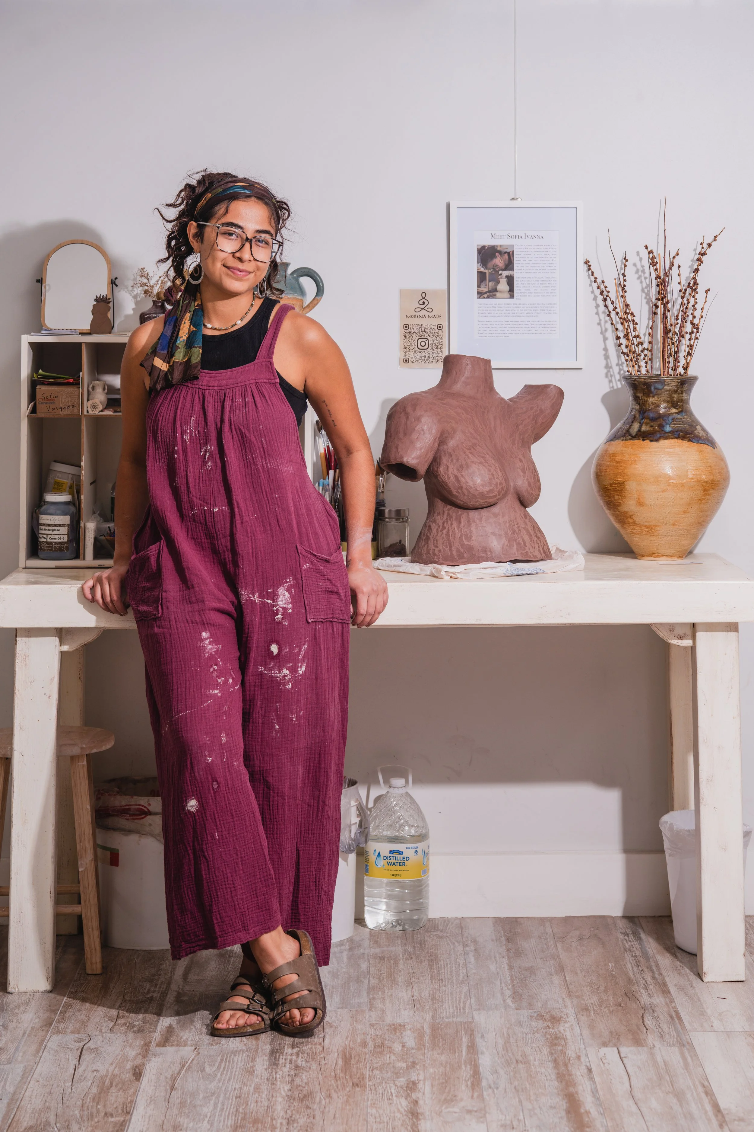 A woman in a black tank top beneath magenta overalls, standing in an art studio with a sculpture of a human torso on the table behind her.