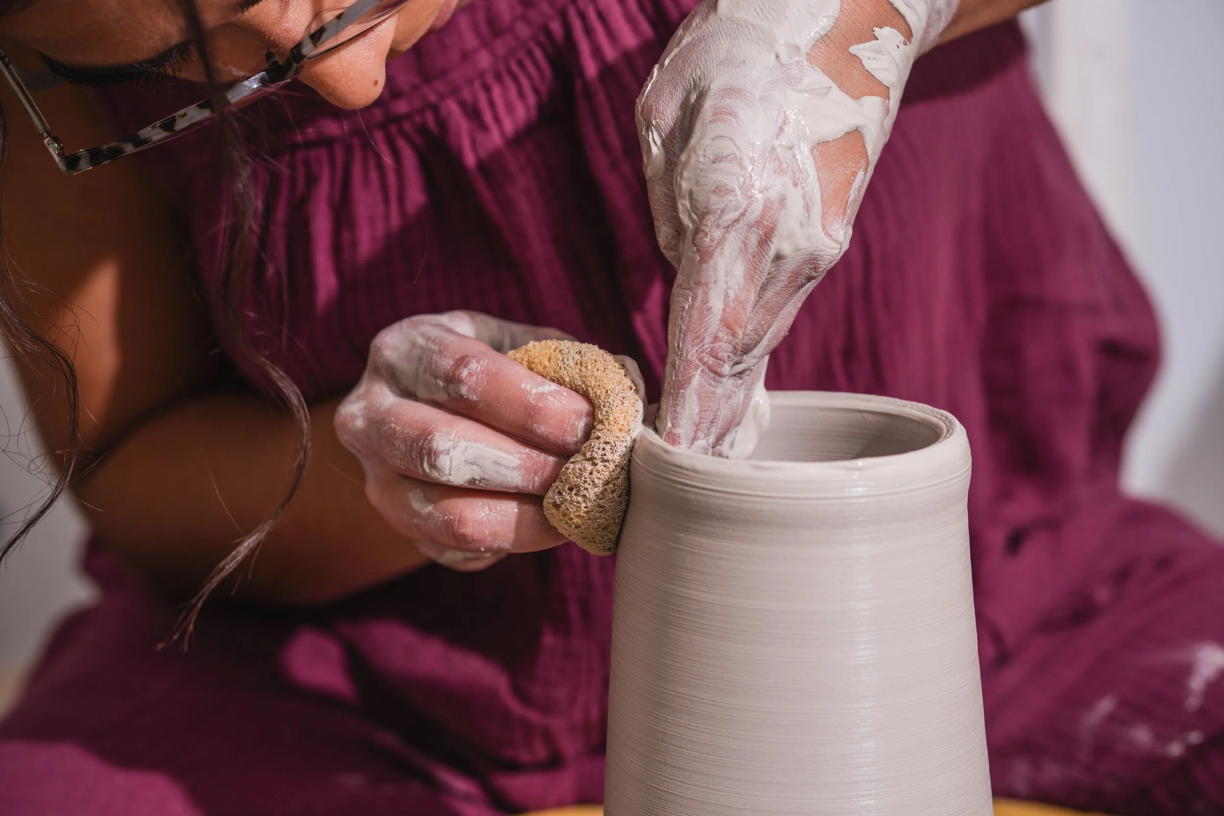 A person shaping a ceramic vase on a pottery wheel with a sponge, wearing a pinkish-purple garment and using a sponge on their fingertips.