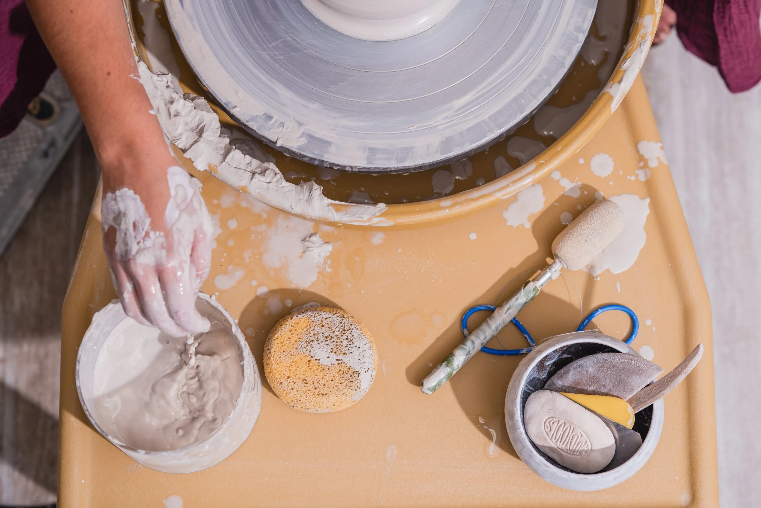 Overhead view of a cake decorator's workstation with a turntable, a bowl of grey icing, a wet sponge, a foam paint roller, a small bowl with spatulas, and spatulas of different sizes and shapes, surrounded by smears of icing and crumbs on a yellow ta