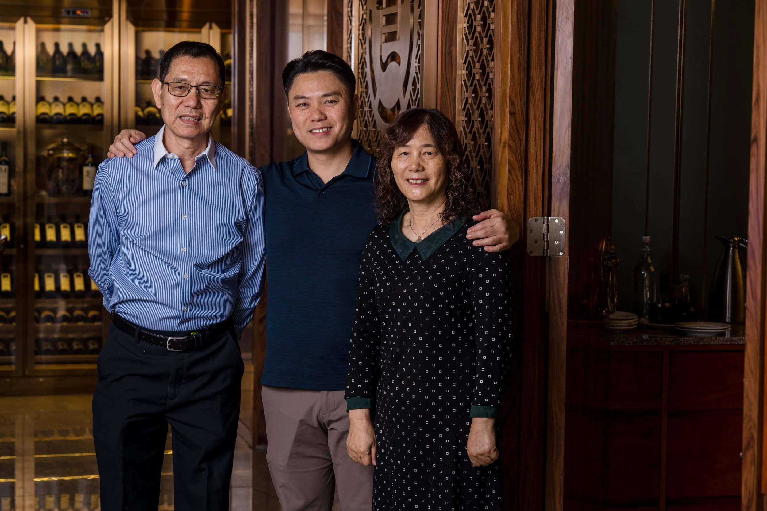 Three people standing together indoors, smiling, with a wooden wall and a cabinet with wine bottles in the background.