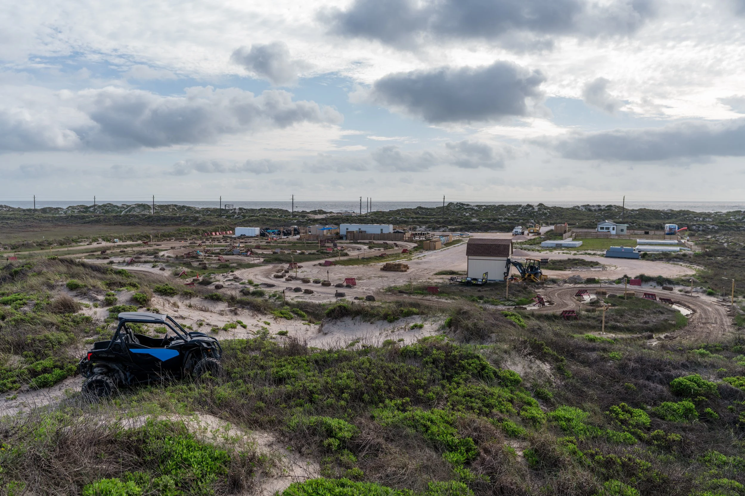 A construction site in a coastal area with a dirt road, construction equipment, small buildings, and a black off-road vehicle in green shrubbery in the foreground. Overcast sky with partly cloudy weather.