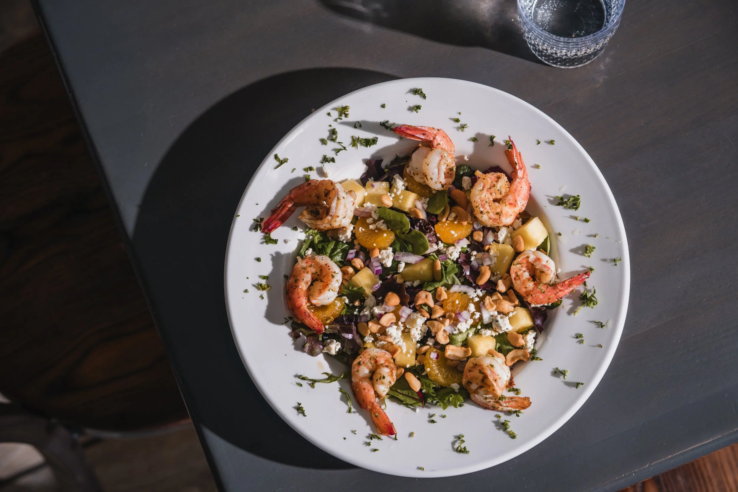 Shrimp salad with mixed greens, pineapple, mandarin oranges, chopped nuts, and crumbled cheese on a white plate.