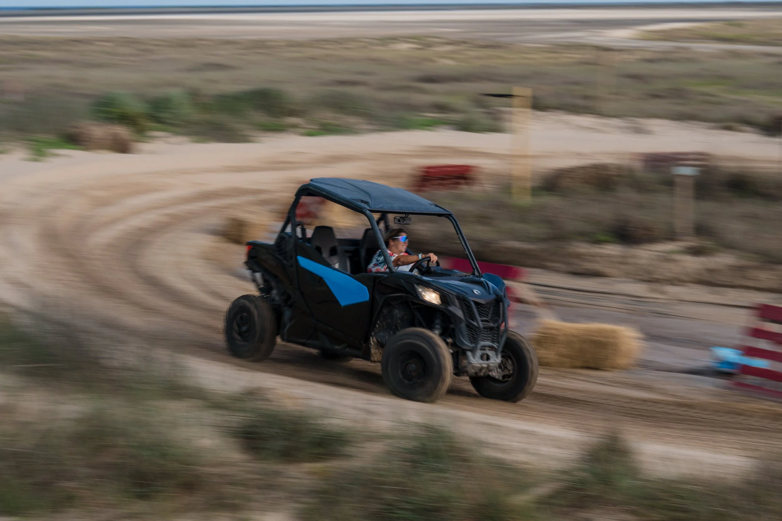 A woman driving a black and blue off-road side-by-side vehicle on a dirt track with hay bales and barriers.