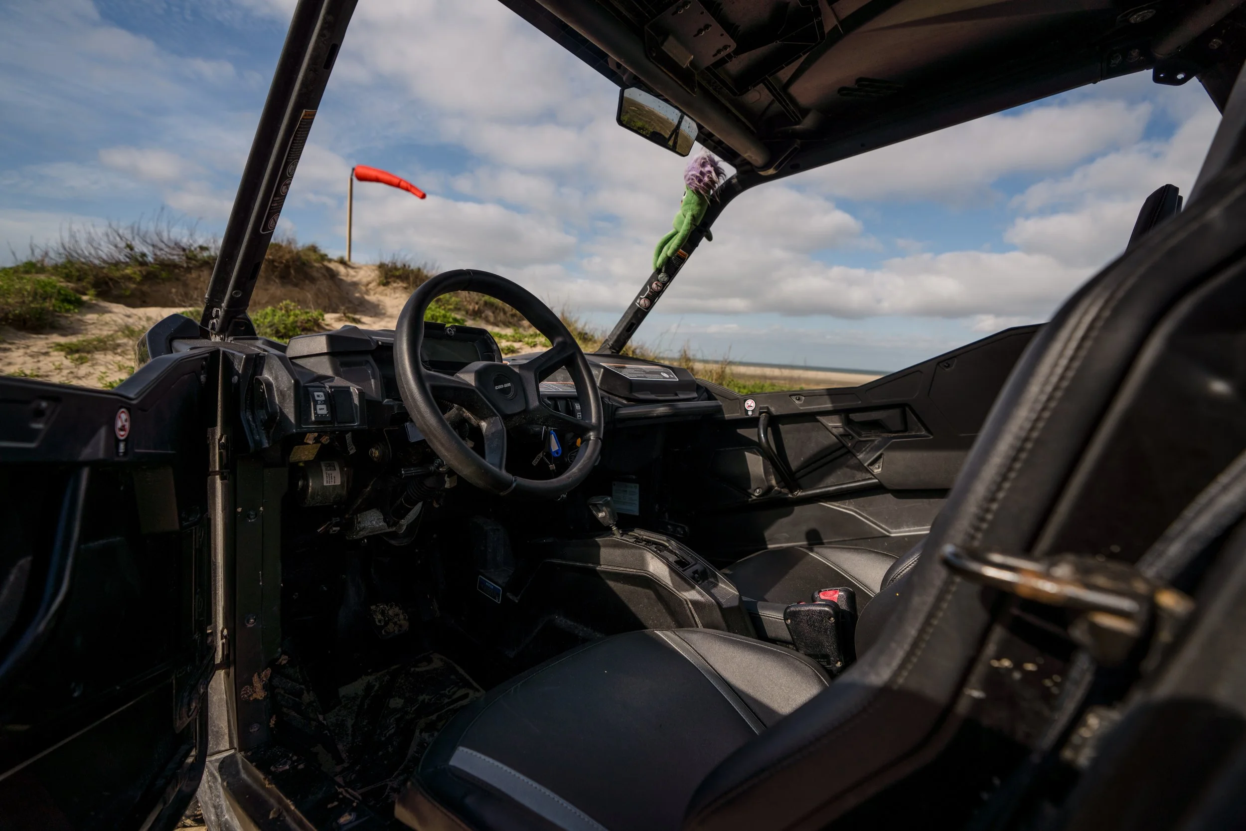 The interior of a black off-road utility vehicle parked on sandy terrain, with a cloudy sky and a windsock visible outside.