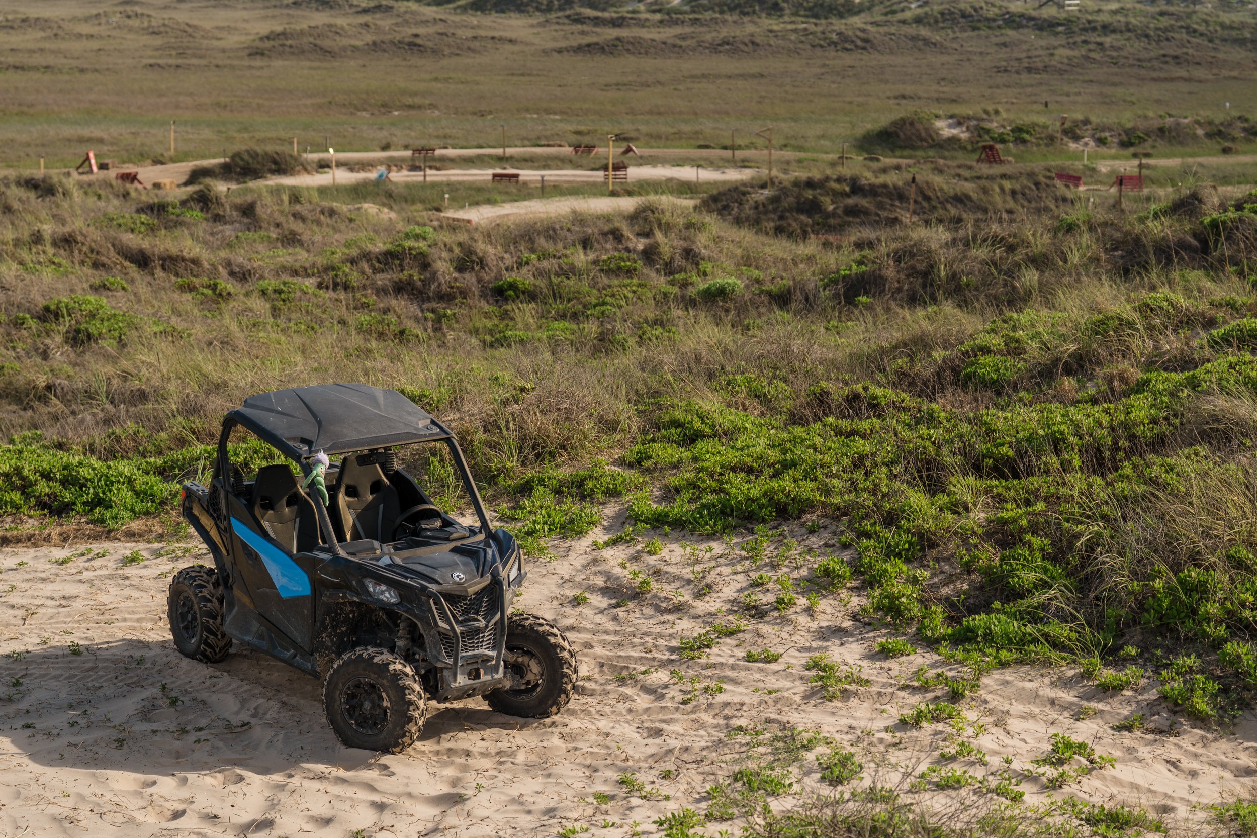 A black off-road utility vehicle parked on sandy terrain with green bushes, overlooking a hilly landscape with a walking trail and benches.
