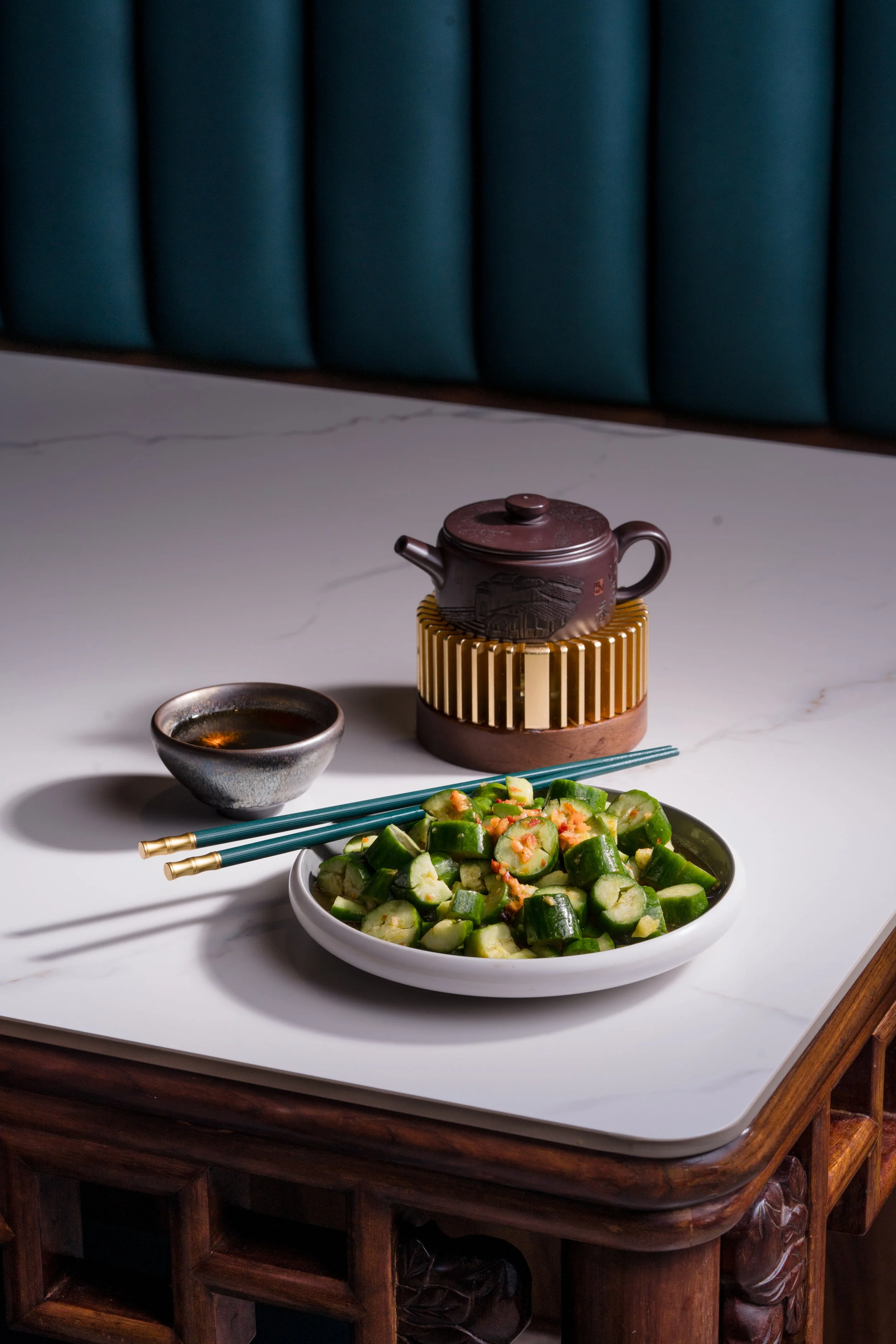 A white plate filled with chopped cucumbers topped with a garnish, placed on a white marble table. There are teal chopsticks resting on the plate, a small black bowl of sauce nearby, and a traditional teapot on a stand behind the plate. A teal uphols