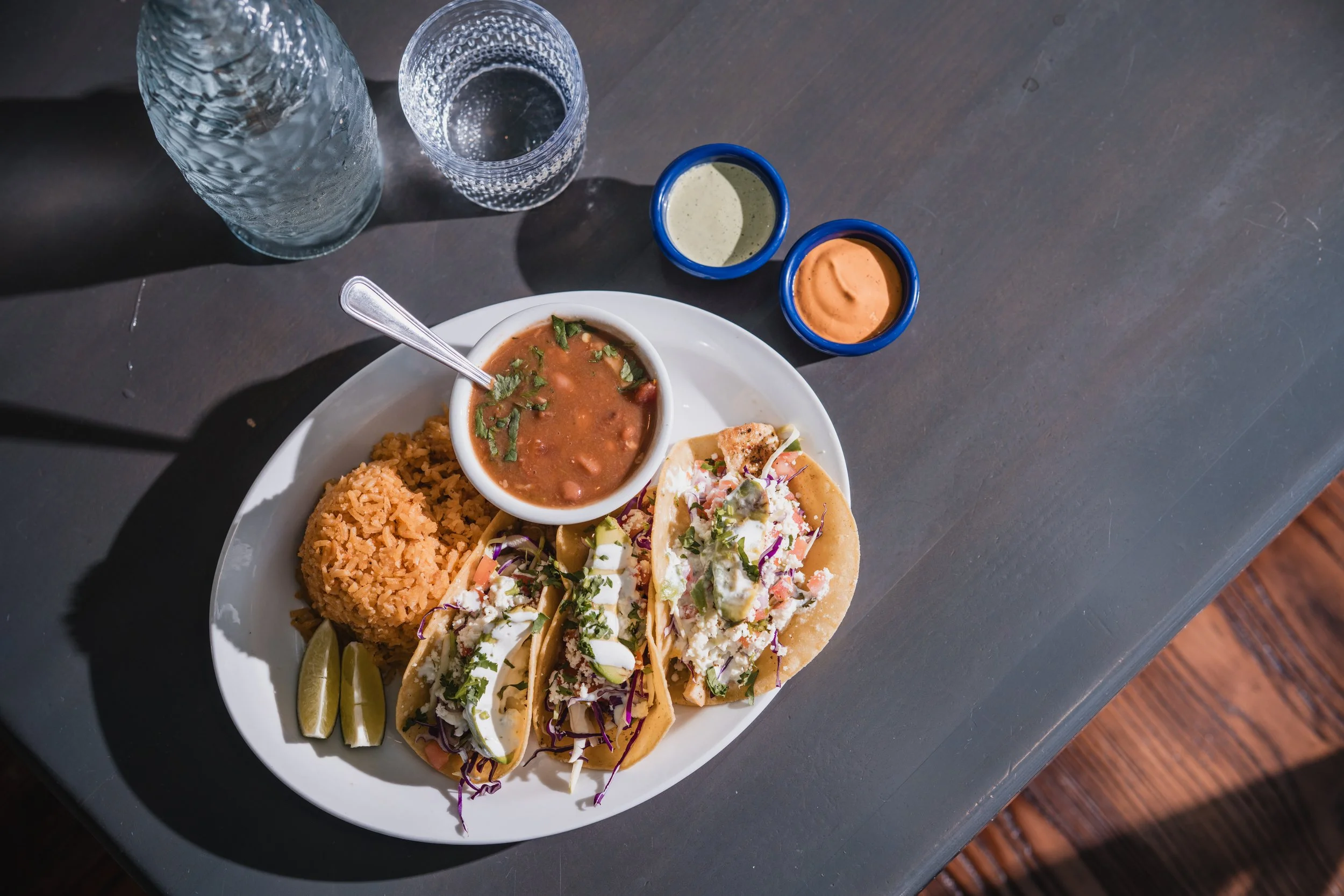 A plate with two tacos topped with shredded cabbage, cilantro, and cheese, a serving of rice, and a bowl of beans. Two small bowls of salsa and a wedge of lime on a dark gray table, with water glasses nearby.