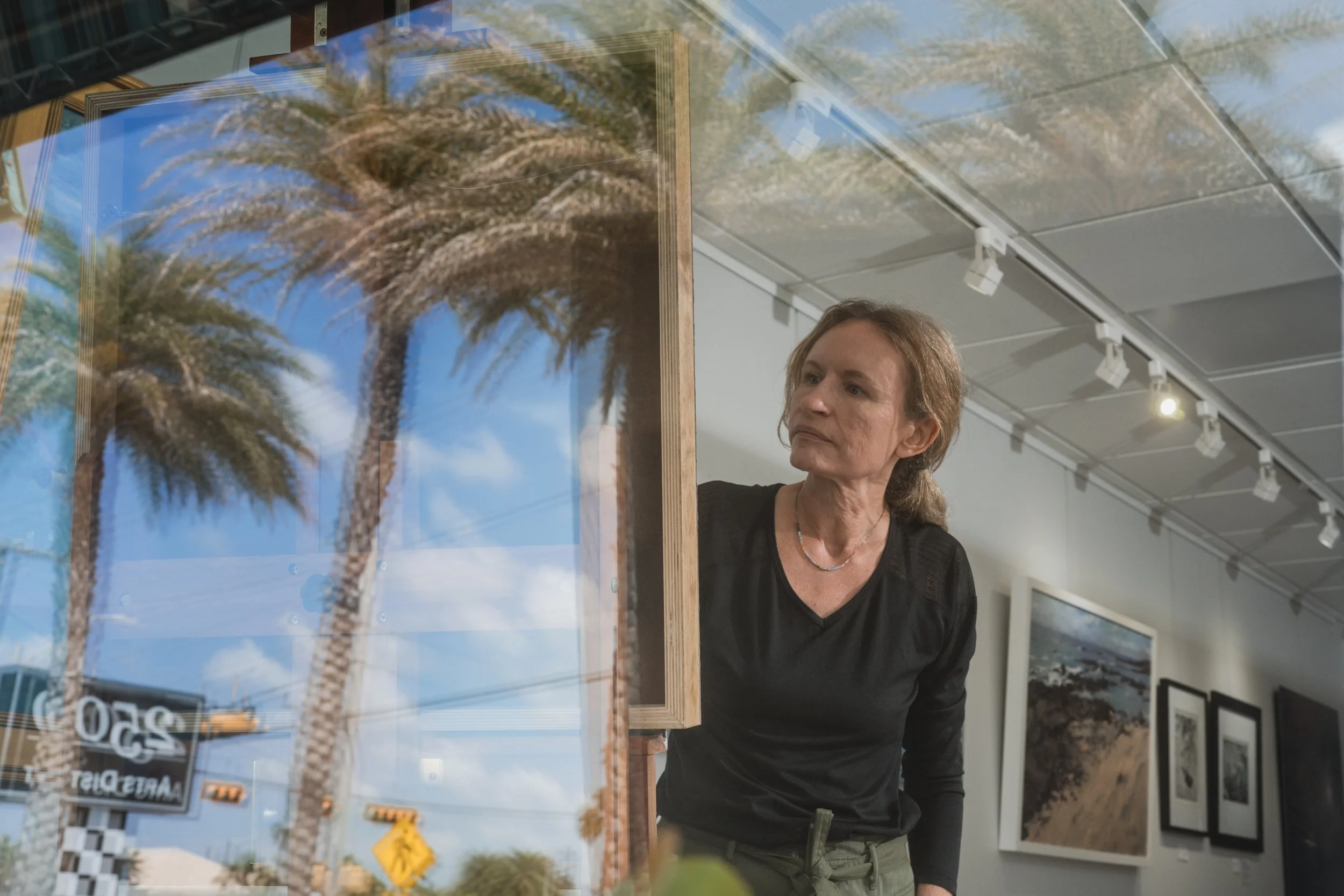 A woman looking thoughtfully at framed artwork of palm trees inside an art gallery, with reflections of palm trees on the glass.