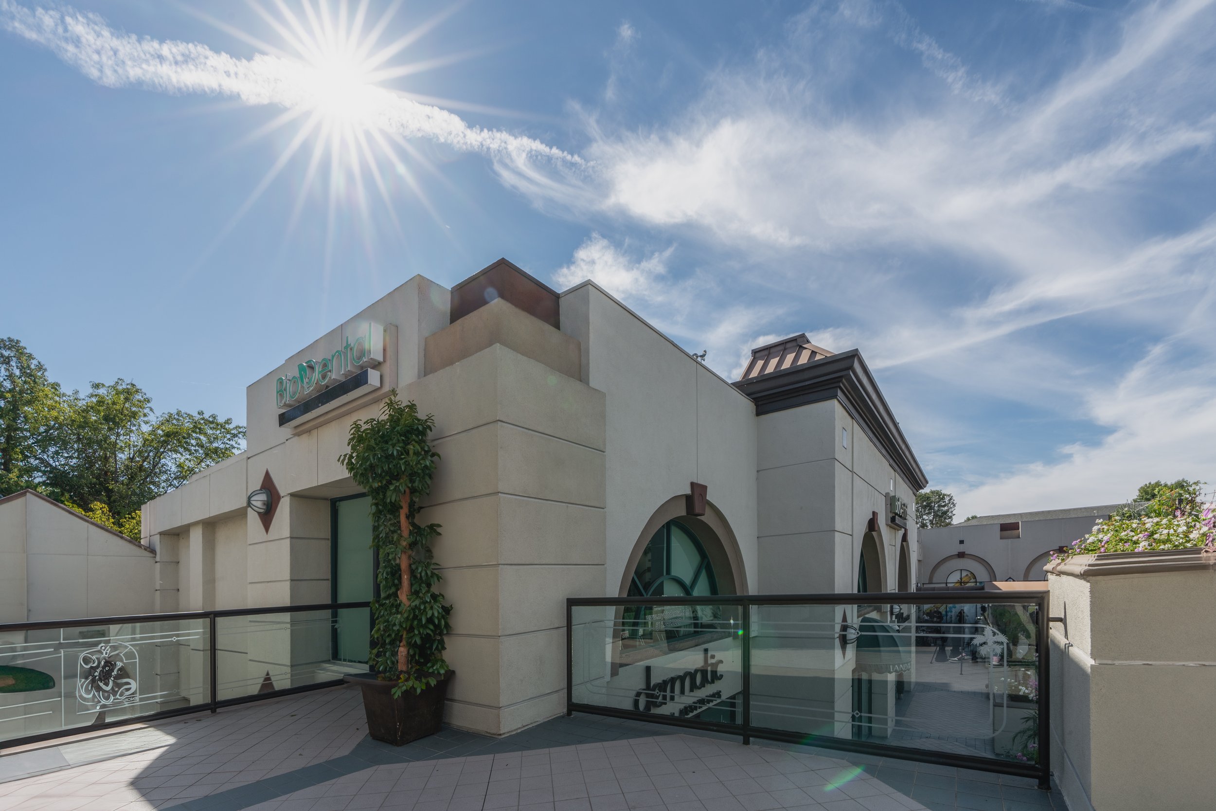 Exterior view of a modern commercial building with the sign 'BioDental' under the bright sun, blue sky, and some clouds, with a potted plant on the balcony.
