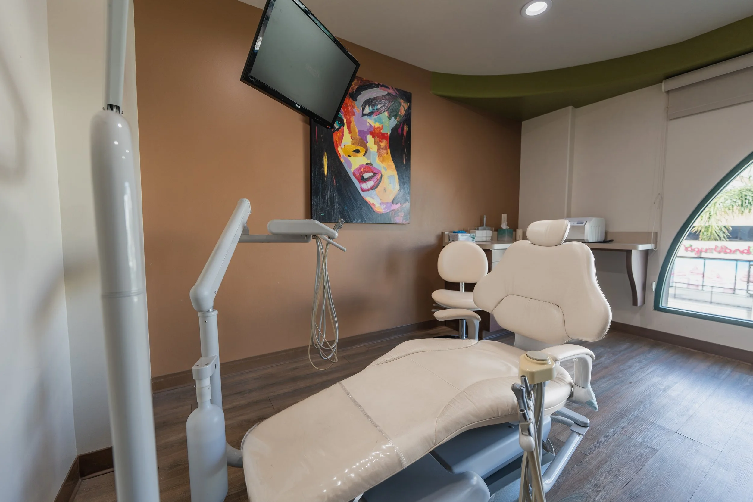 Dental or medical examination room with a reclining chair, a mounted flat-screen monitor, a colorful portrait on the wall, and a small cabinet with medical supplies near a window.
