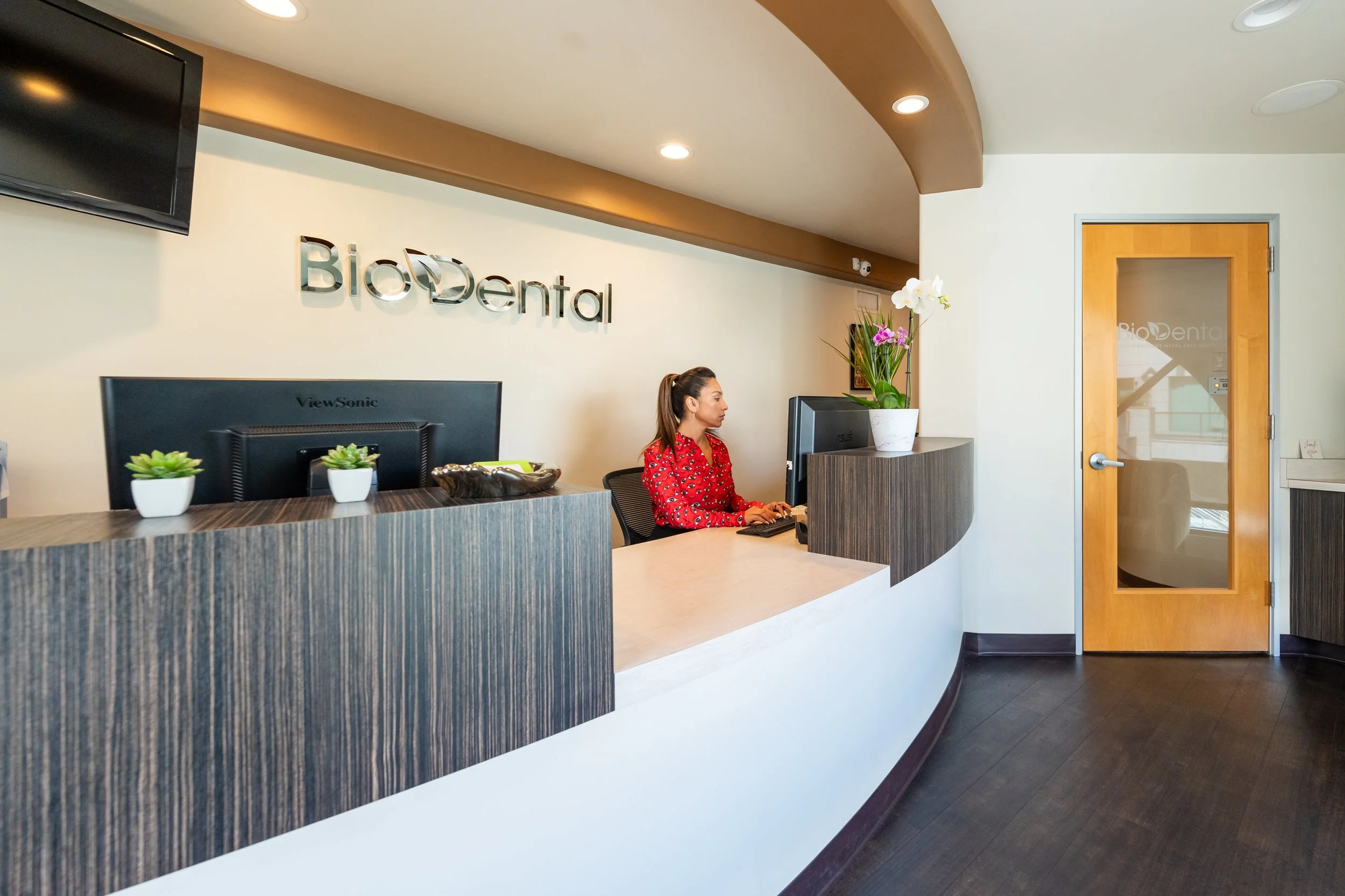 Reception area of BioDental dental clinic with a woman sitting at the counter using a computer, white walls, a wooden door with a glass panel, and plants on the counter.