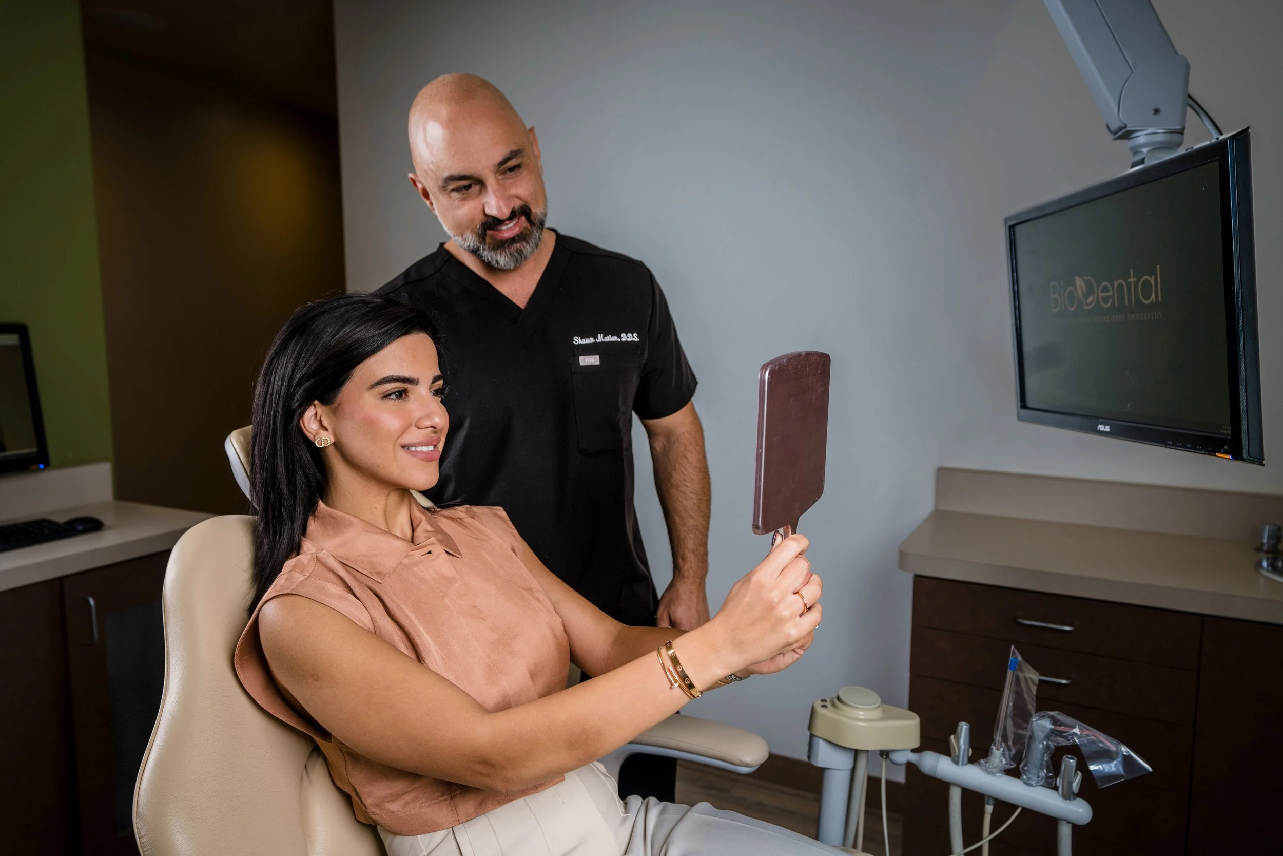 A woman sitting in a dental chair holding a mirror, with a dentist looking on in a dental office.