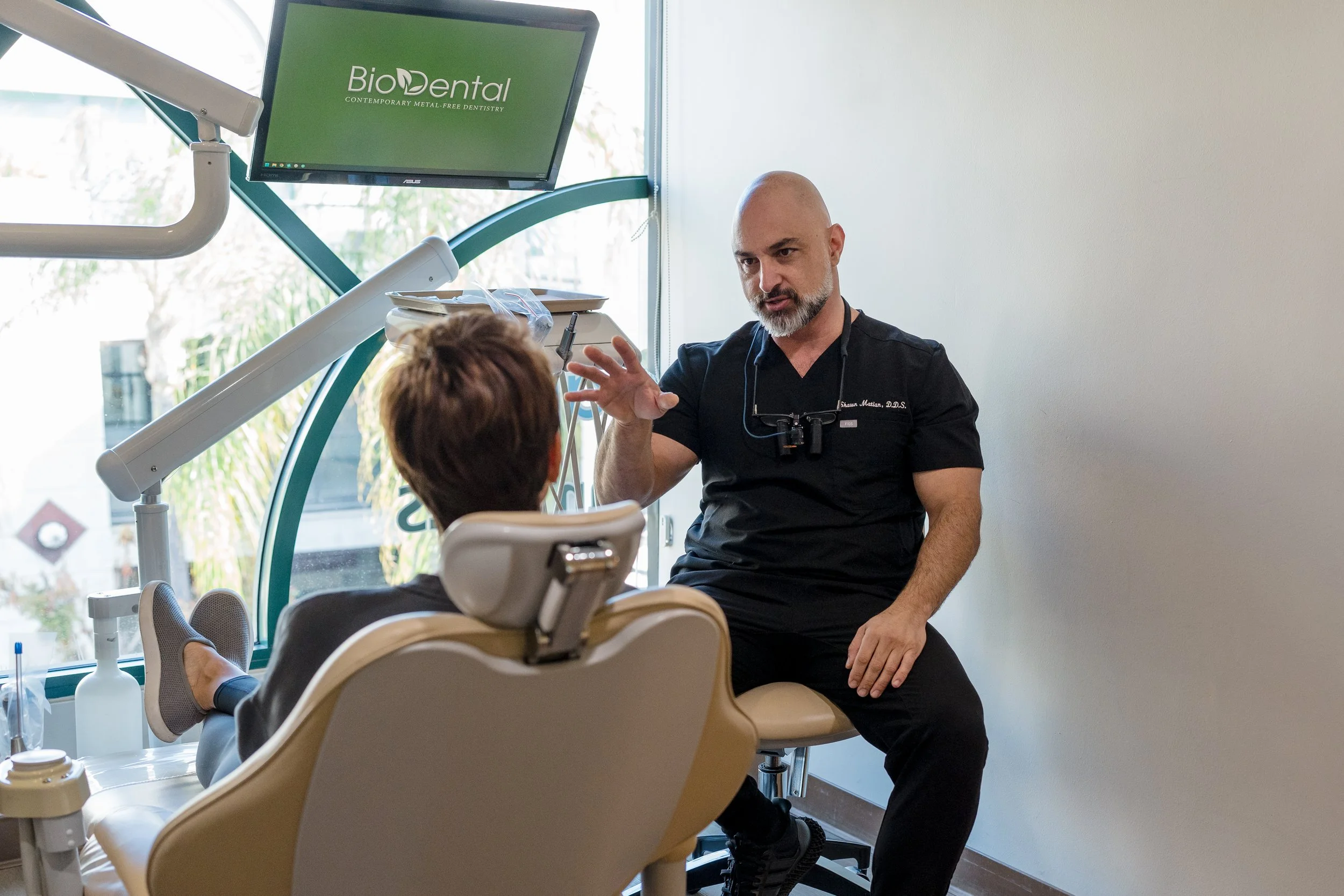 A dentist talking to a patient seated in a dental chair at BioDental clinic, with dental tools and a monitor in the background.