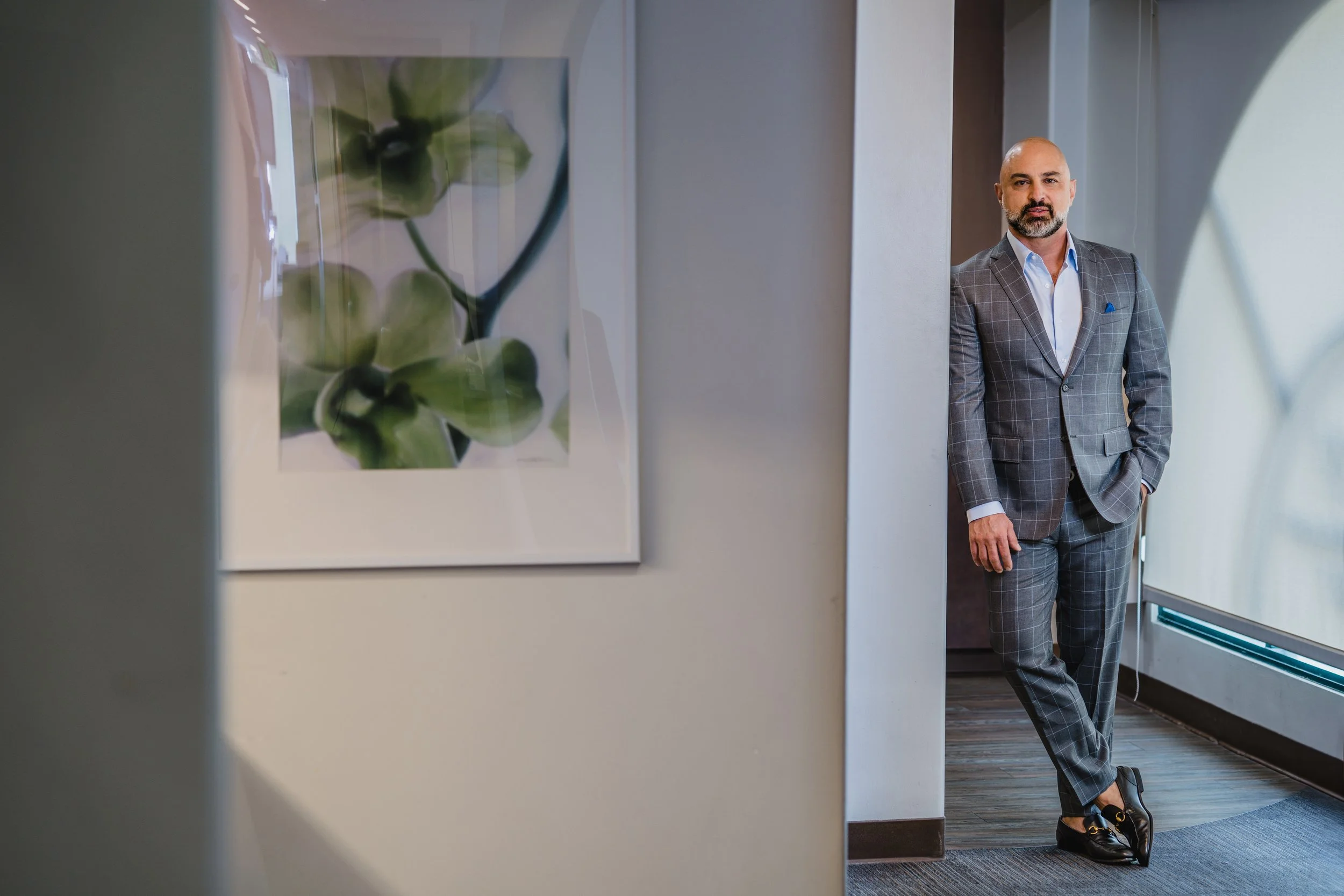 A man in a gray plaid suit standing in a hallway near a large window with a view outside, next to a framed botanical photograph of green orchids on the wall.