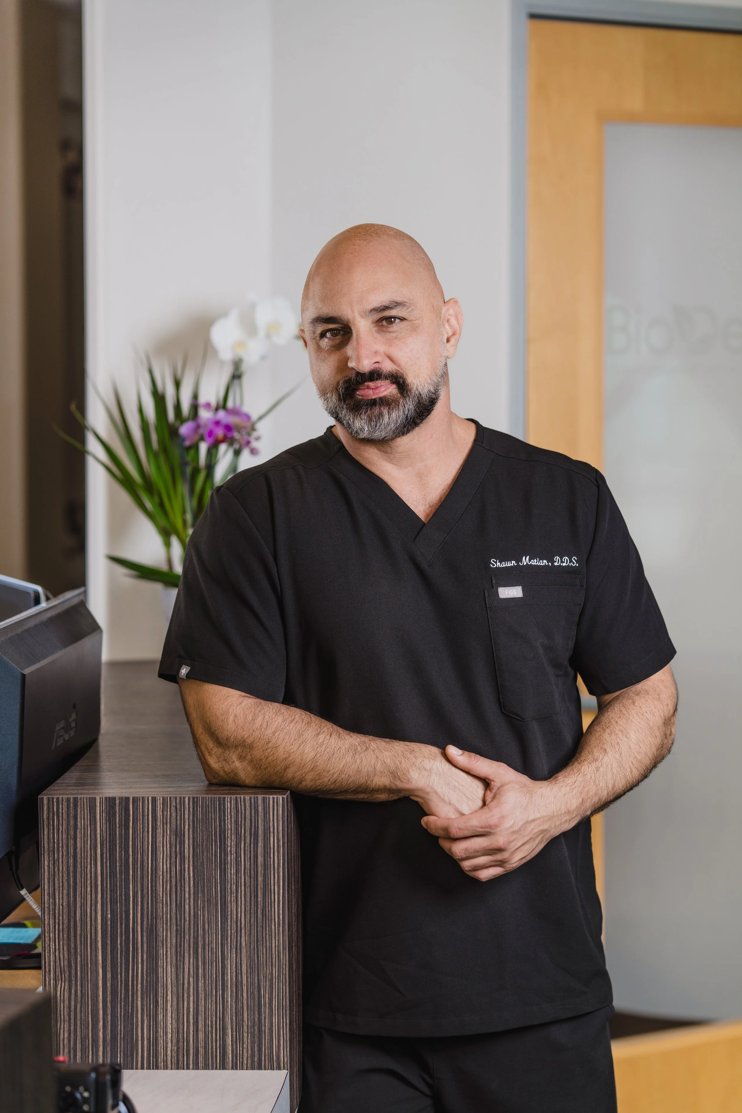 A male dentist with a beard and shaved head standing at a reception desk, dressed in black scrubs, in a modern dental office with a pot of orchids in the background.