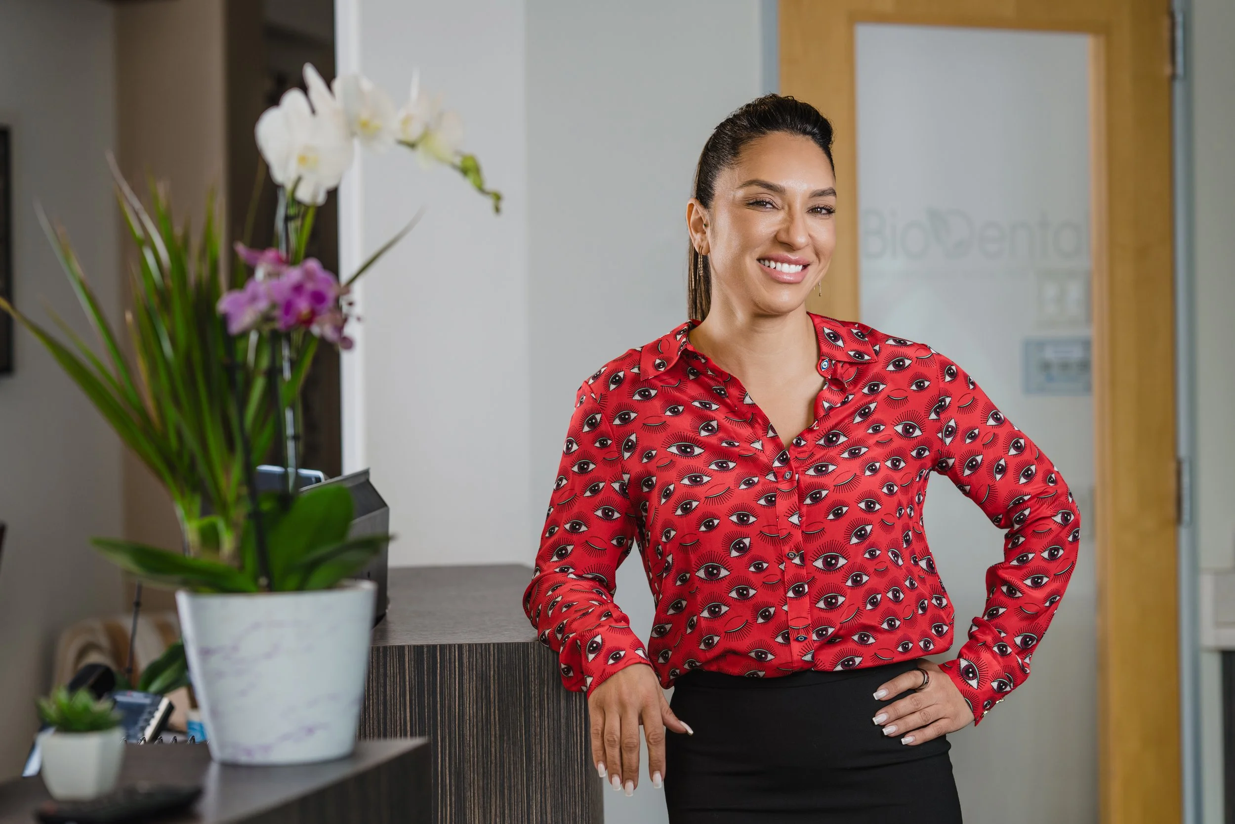 A woman standing in an office lobby, smiling, wearing a red blouse with eye patterns, and a black skirt, with a potted orchid plant nearby.