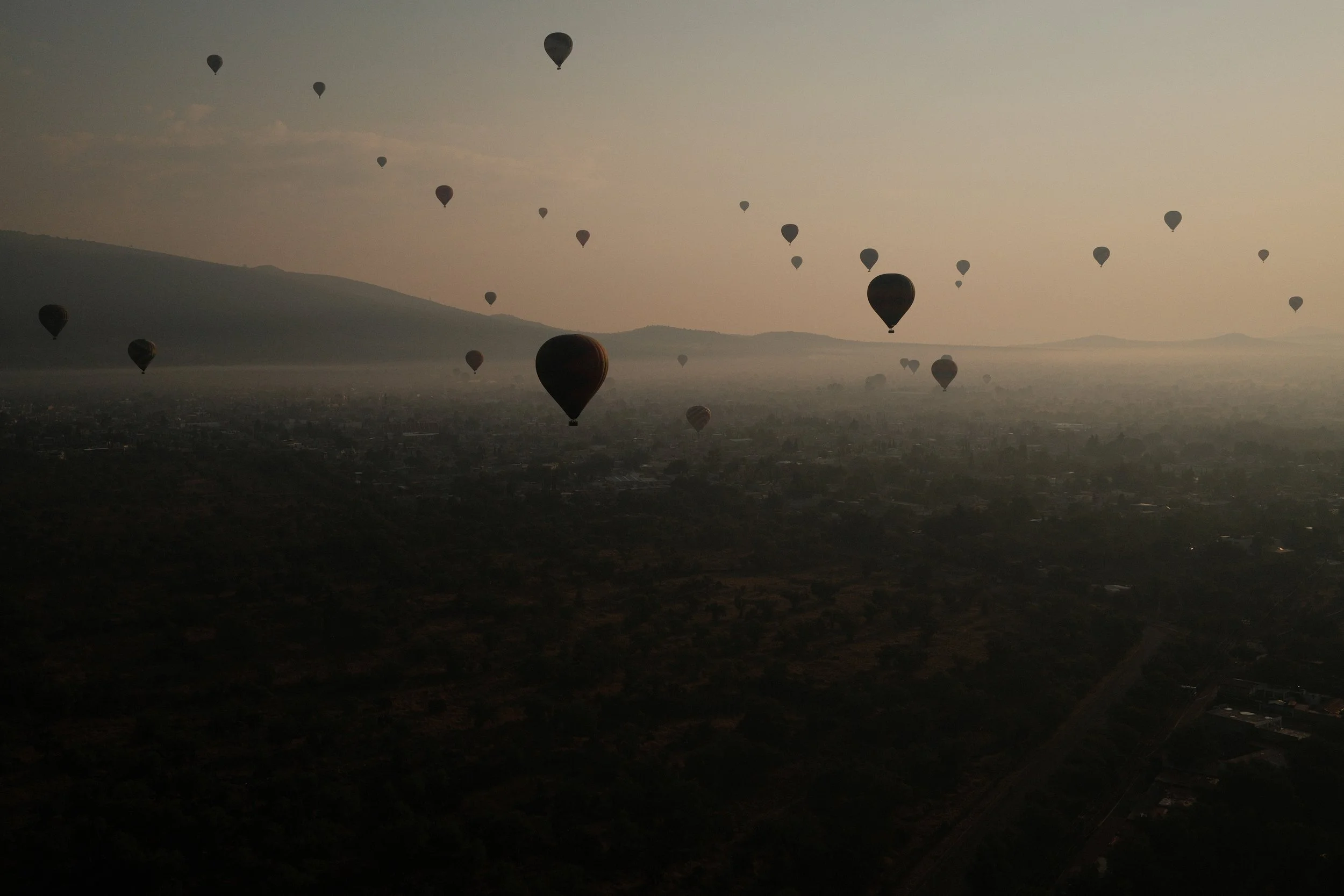 A sky filled with numerous hot air balloons floating over a landscape at sunrise or sunset, with distant hills and a hazy sky.