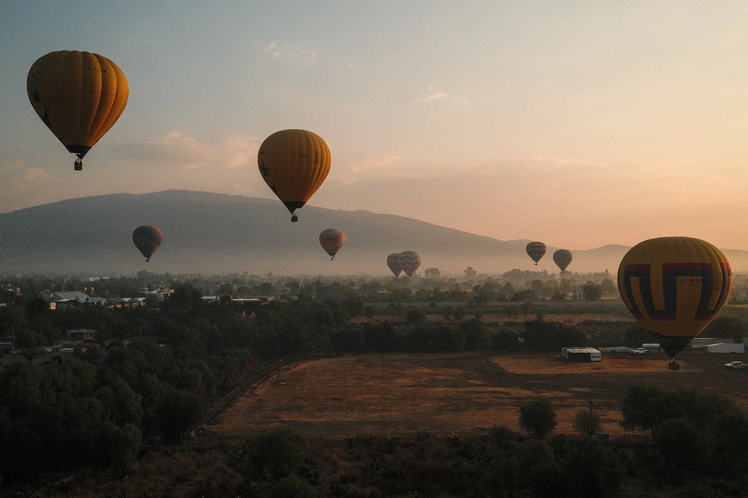 Multiple hot air balloons floating over a landscape at sunrise or sunset, with hills in the background.