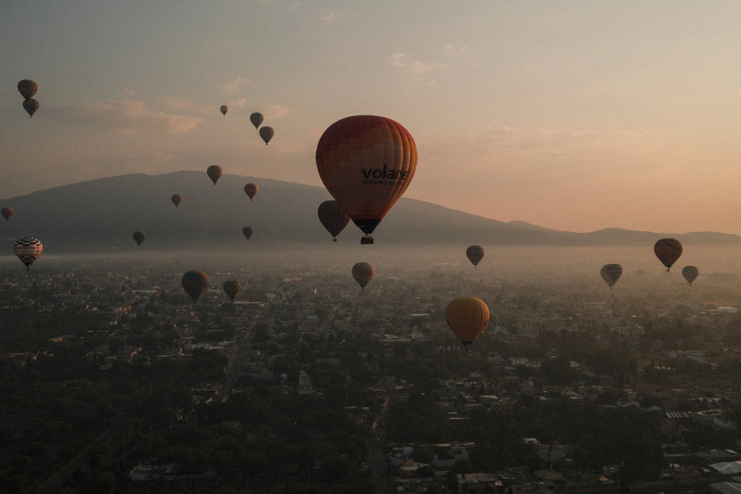 Multiple hot air balloons floating over a city at sunrise or sunset with mountains in the background.