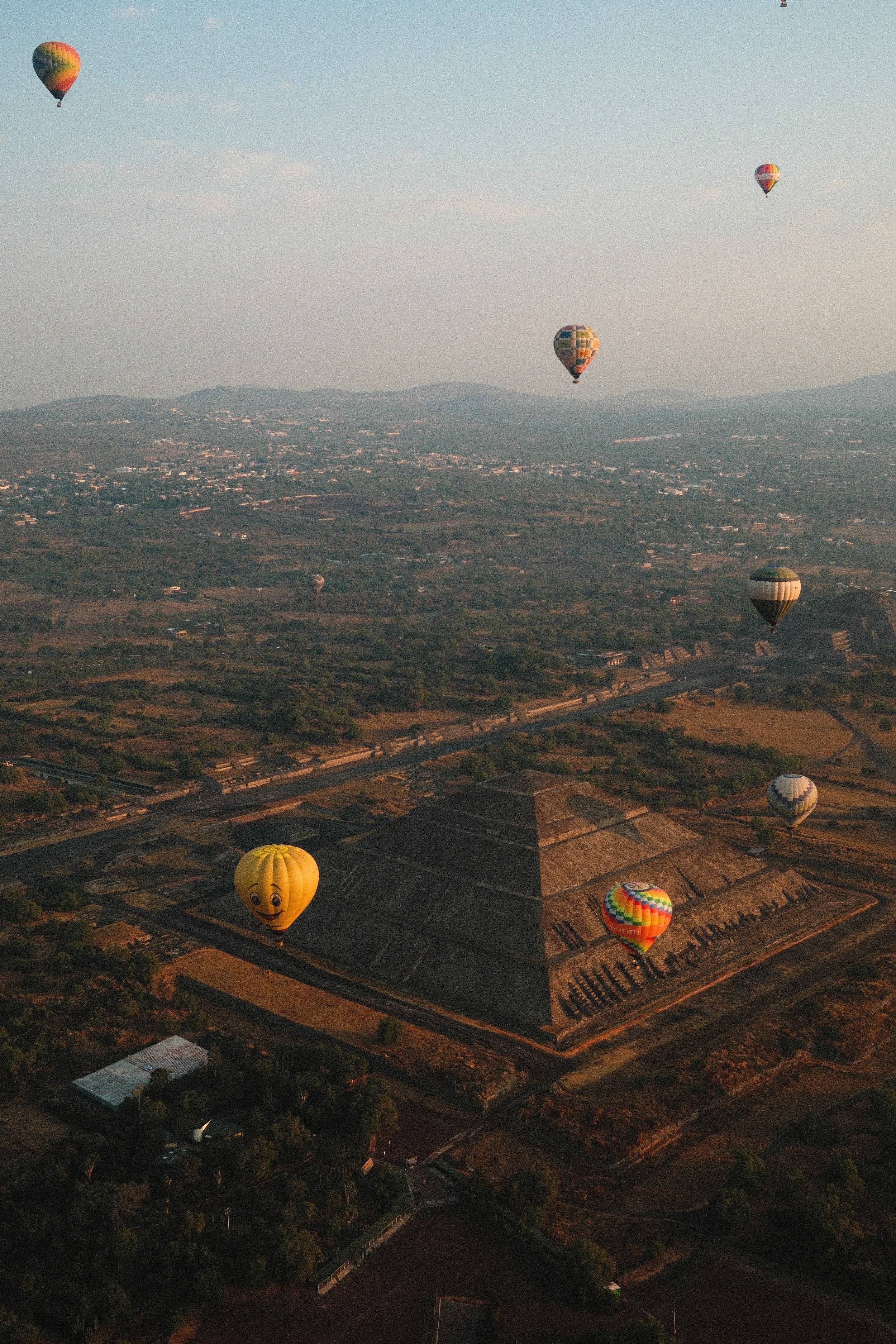 Several colorful hot air balloons floating above the pyramids of Teotihuacan in Mexico during sunrise or sunset.