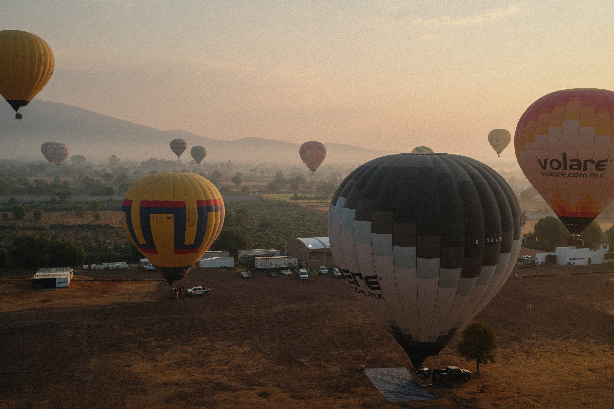 Multiple hot air balloons floating in the sky during sunrise or sunset over a rural landscape with fields, trees, and mountains in the background.