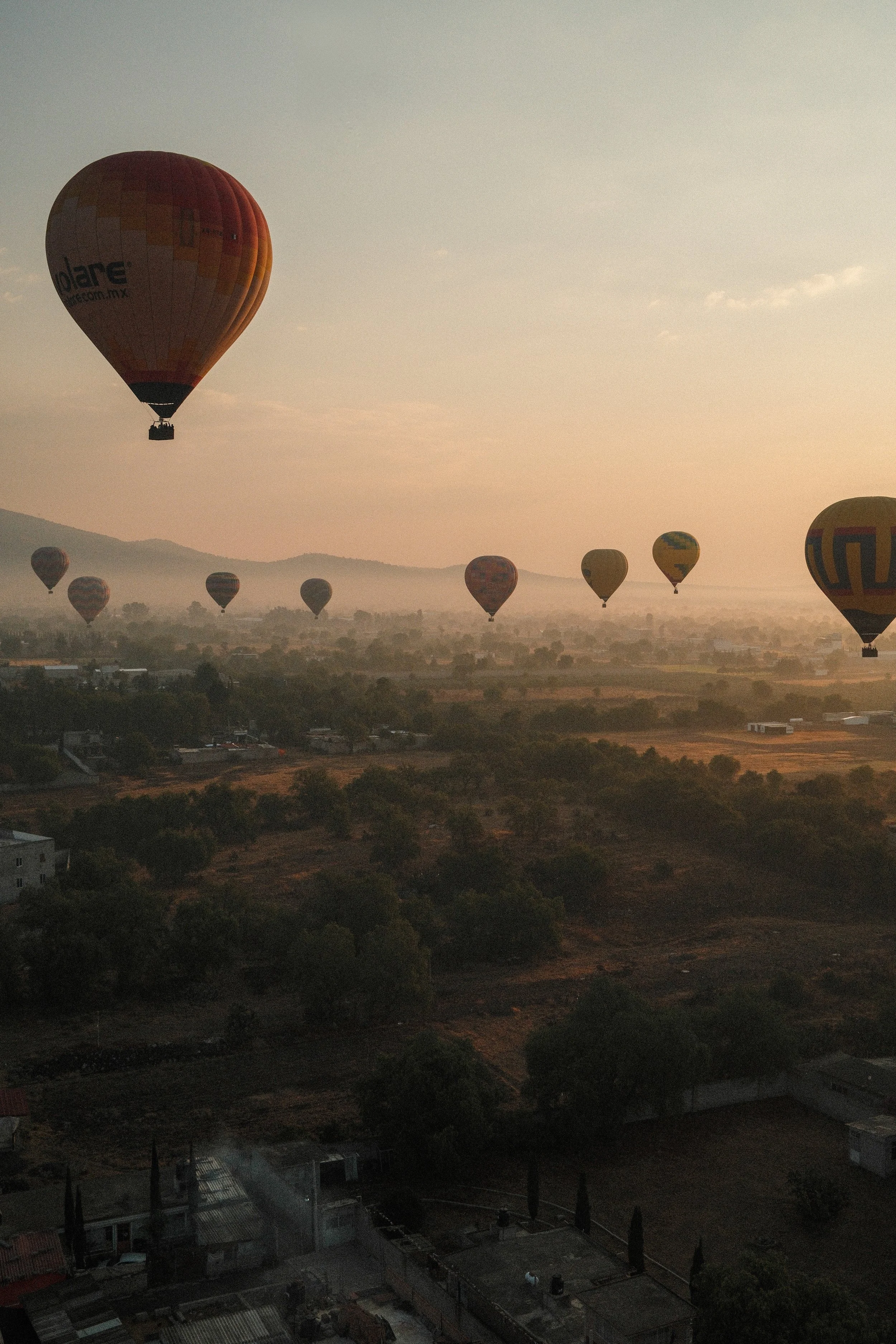 Multiple colorful hot air balloons floating over a rural landscape at sunrise or sunset, with distant mountains and scattered buildings.