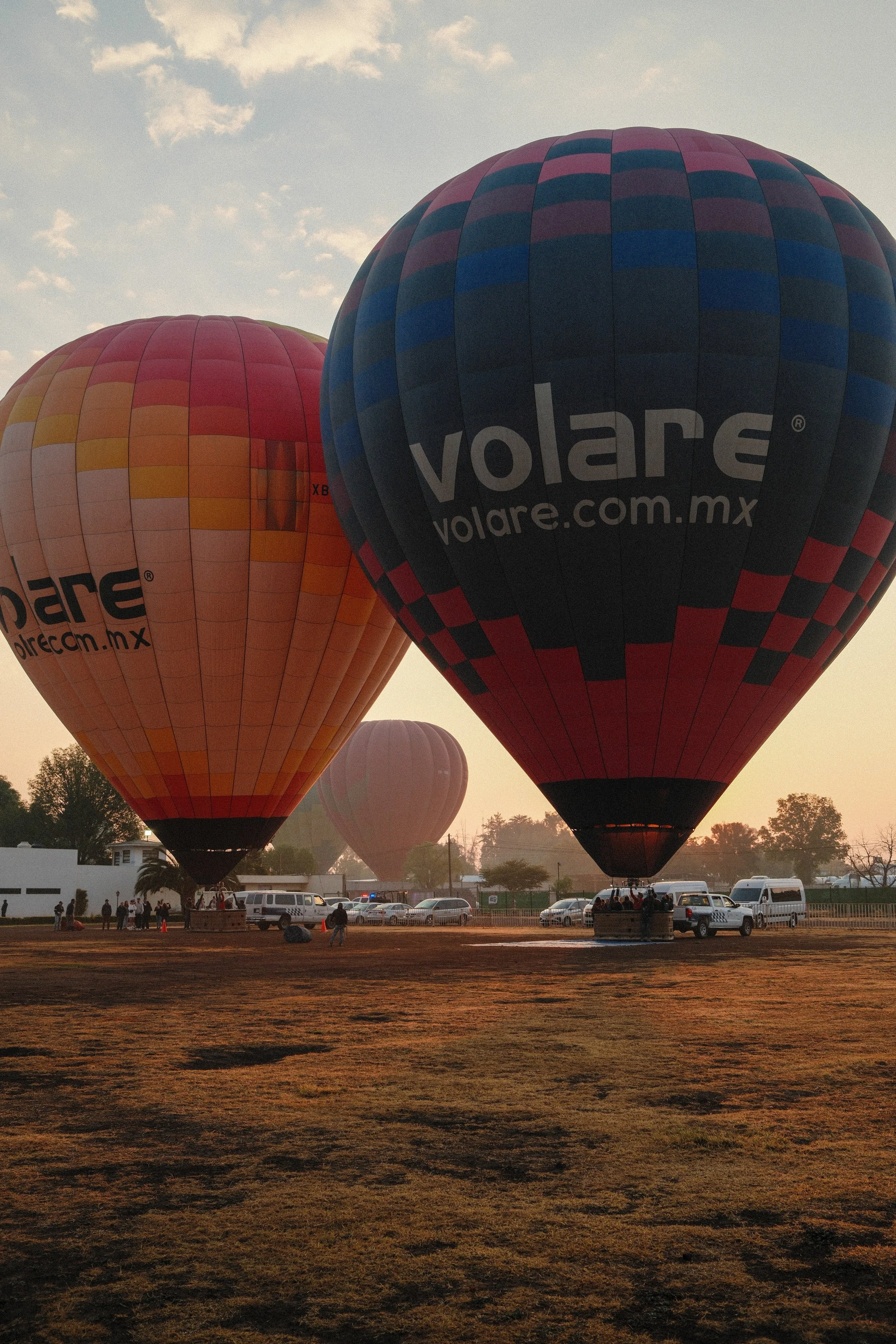 Three hot air balloons floating at sunrise, with people and vehicles on the ground below.