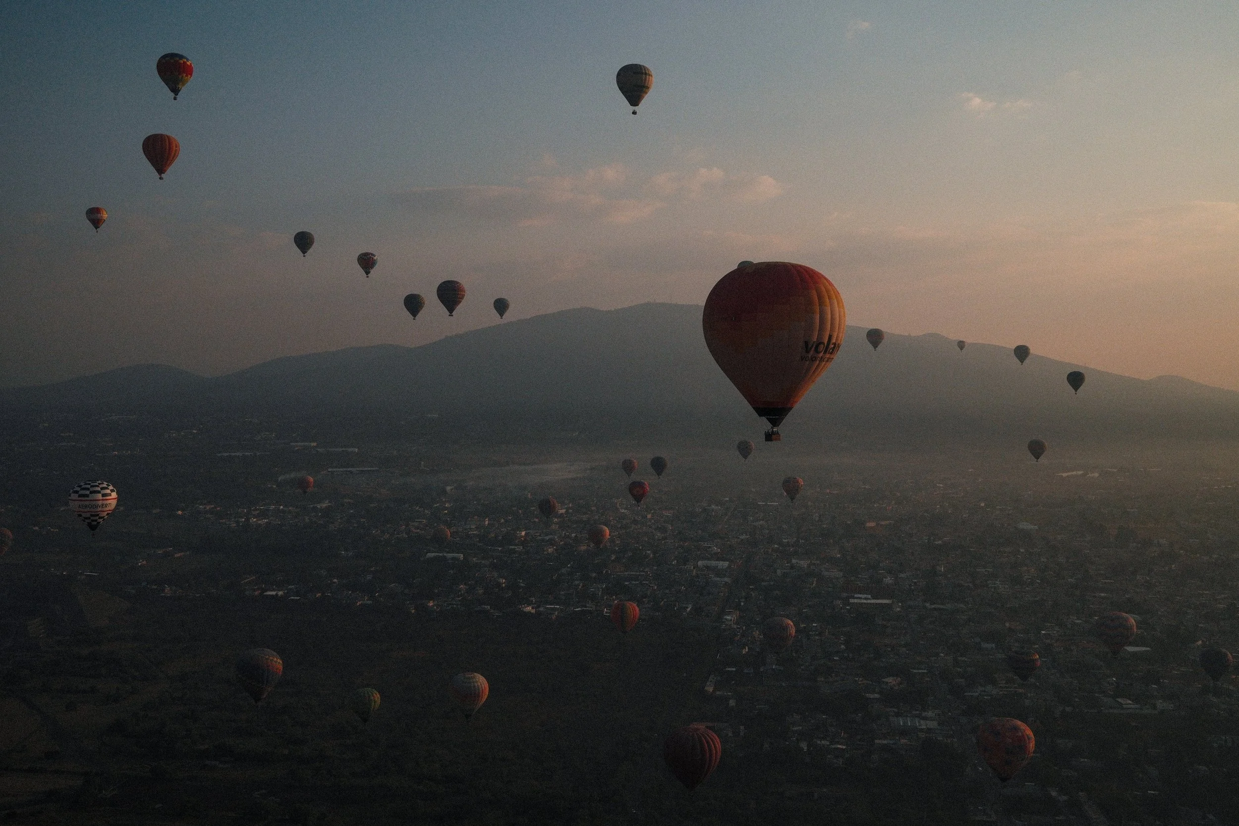 Numerous hot air balloons floating over a city with mountains in the background at dawn or dusk.